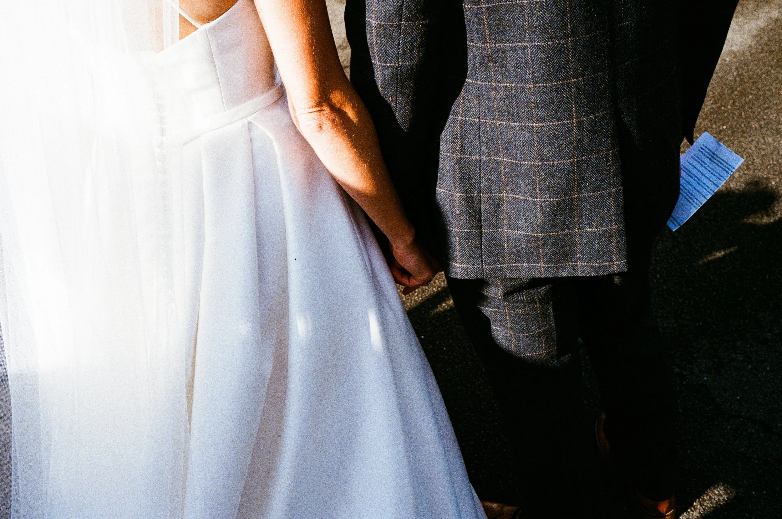 A couple holding hands, dressed in wedding attire, with the woman in a white wedding gown and the man in a plaid suit, standing outdoors.