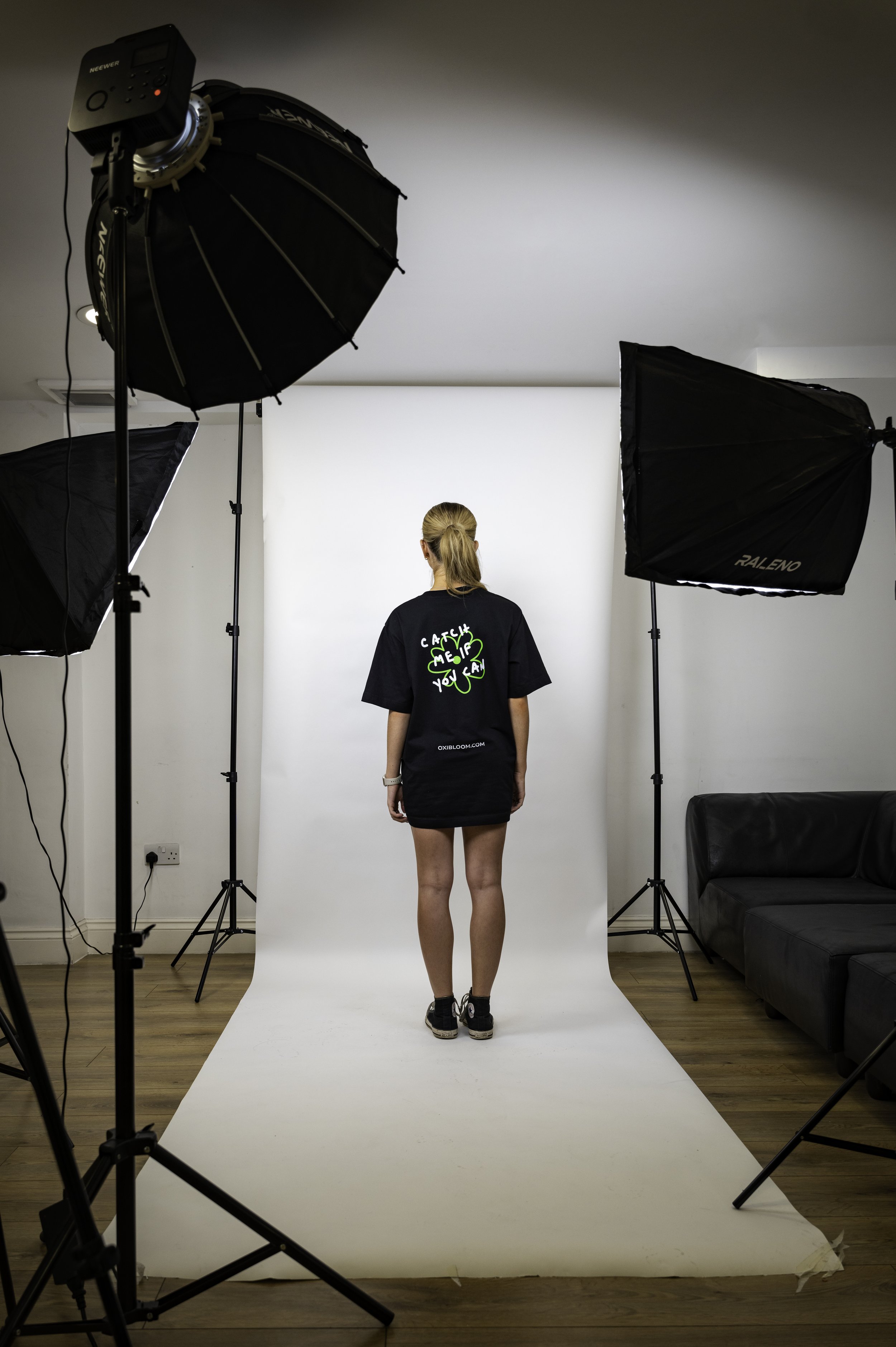 A person with blonde hair in a ponytail standing on a white studio backdrop, surrounded by professional photography lighting equipment in a room with wooden flooring and a black sofa.