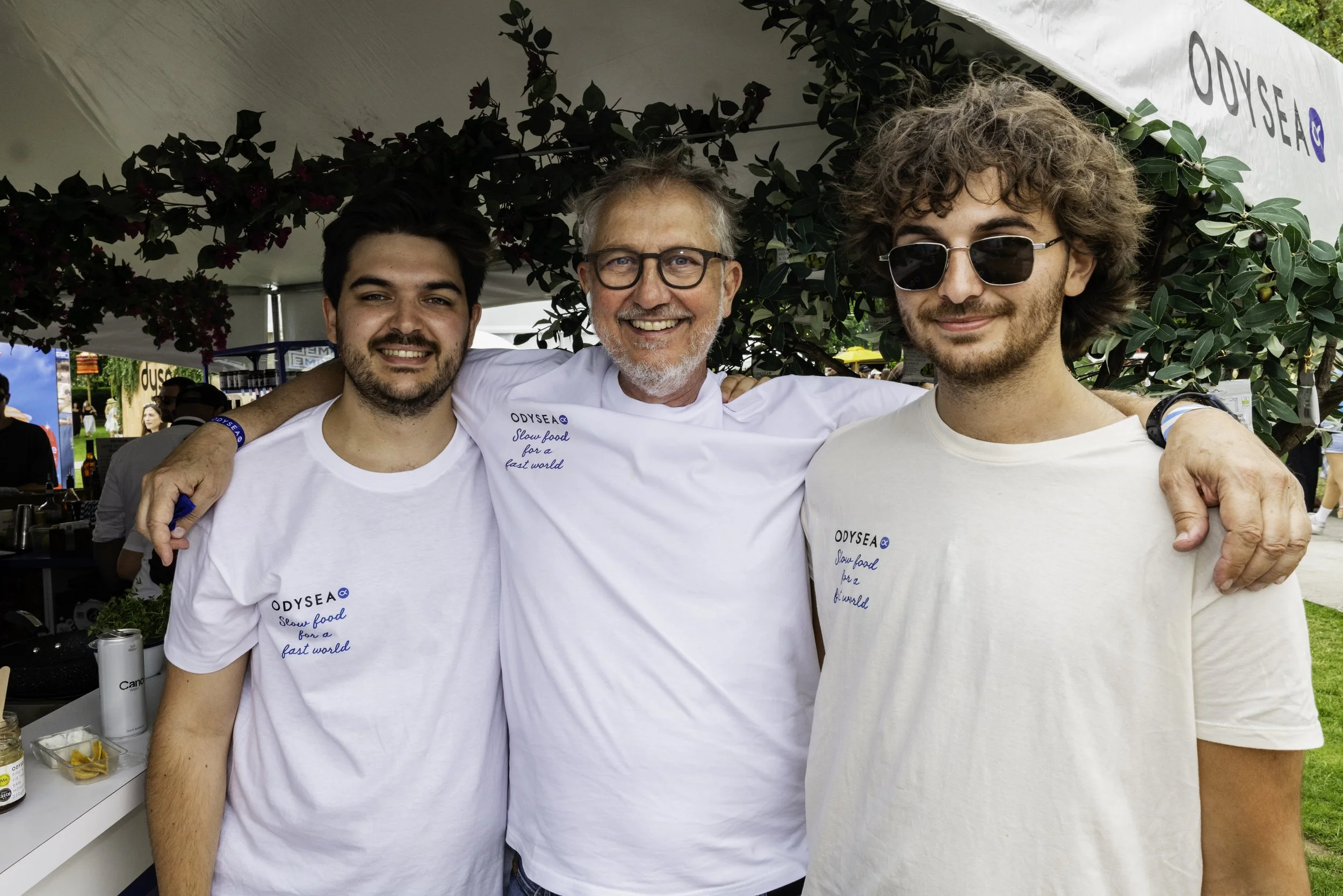 Three men standing under a tent at an outdoor event, smiling and wearing white T-shirts that say 'ODYSEA Slow food for a fast world.' They have their arms around each other.