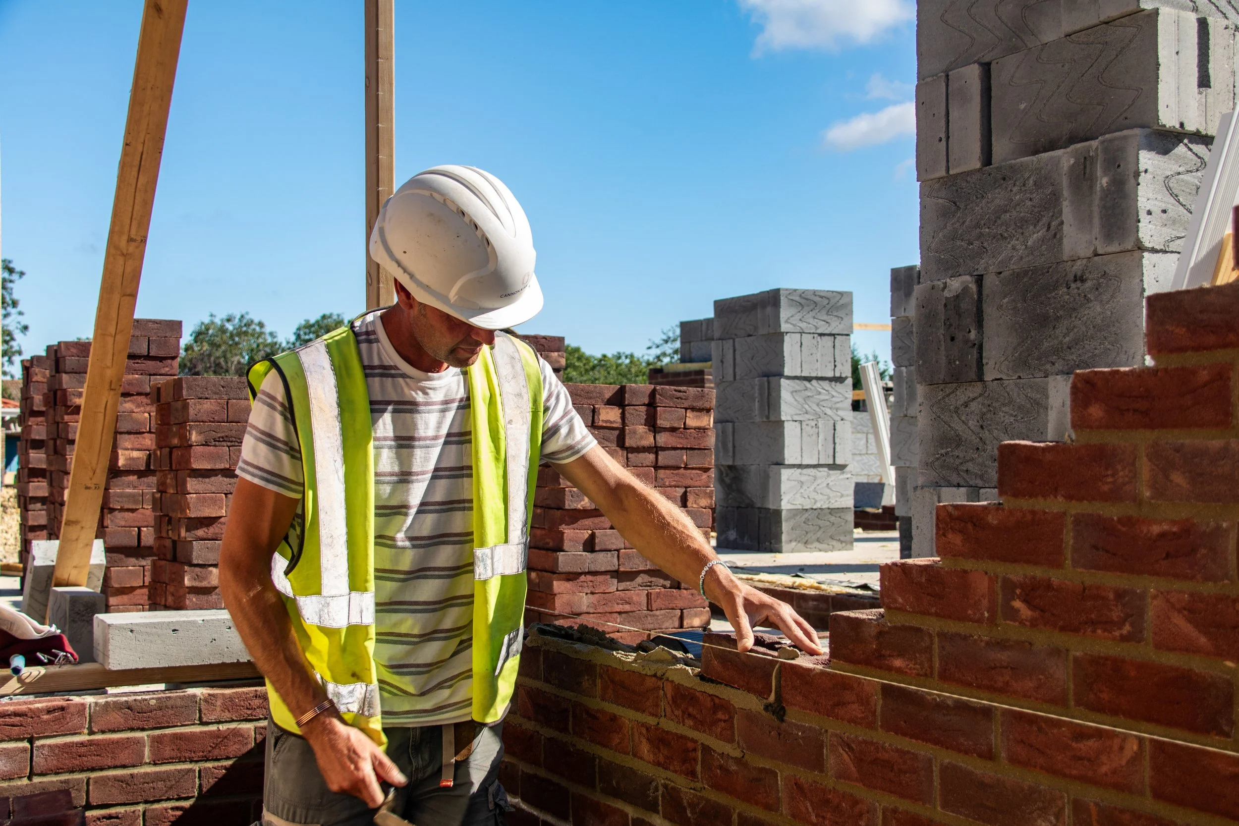 A construction worker wearing a white safety helmet and a high-visibility vest working on a brick wall at a construction site under a clear blue sky.