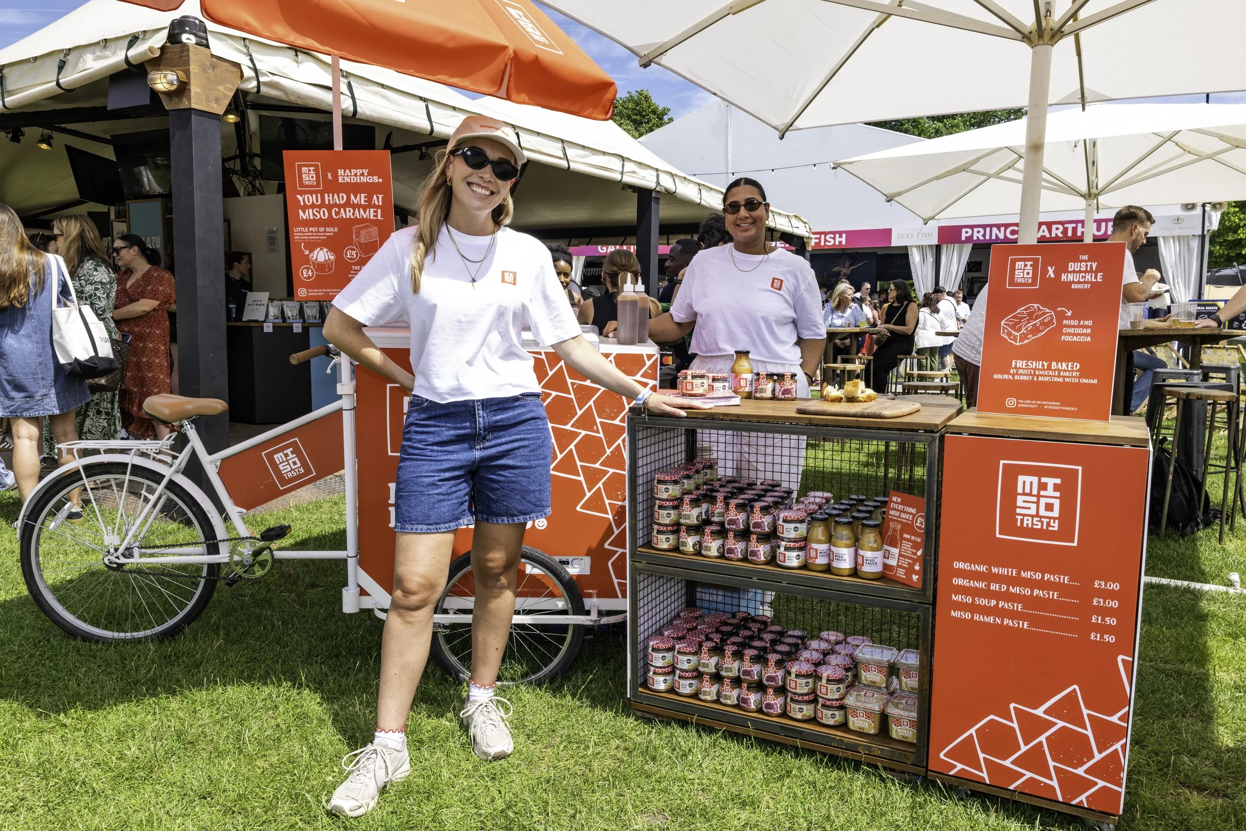 Two smiling women stand at an outdoor food stall selling miso products, with a red sign displaying prices and product descriptions. One woman is wearing sunglasses, a white cap, and a white t-shirt, while the other has dark sunglasses and a white shi