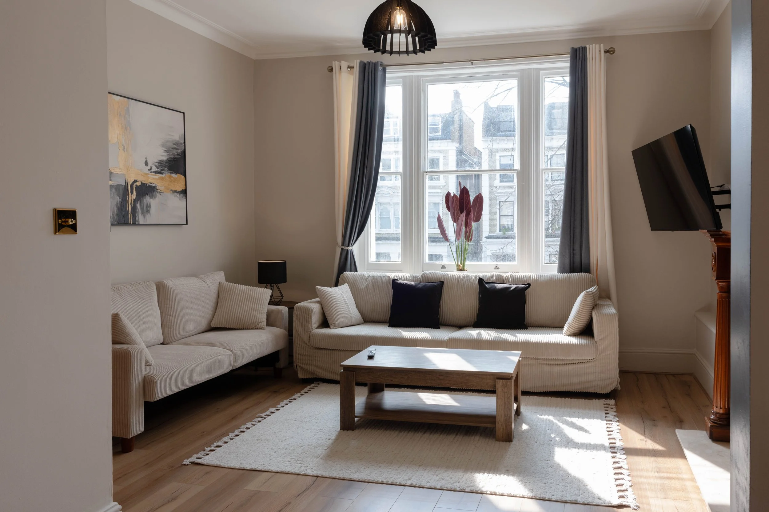 A living room with two beige sofas adorned with beige and black throw pillows, a wooden coffee table, a black table lamp, a large abstract painting on the spacious cream-colored wall, a window with blue and white curtains, a decorative plant on the w