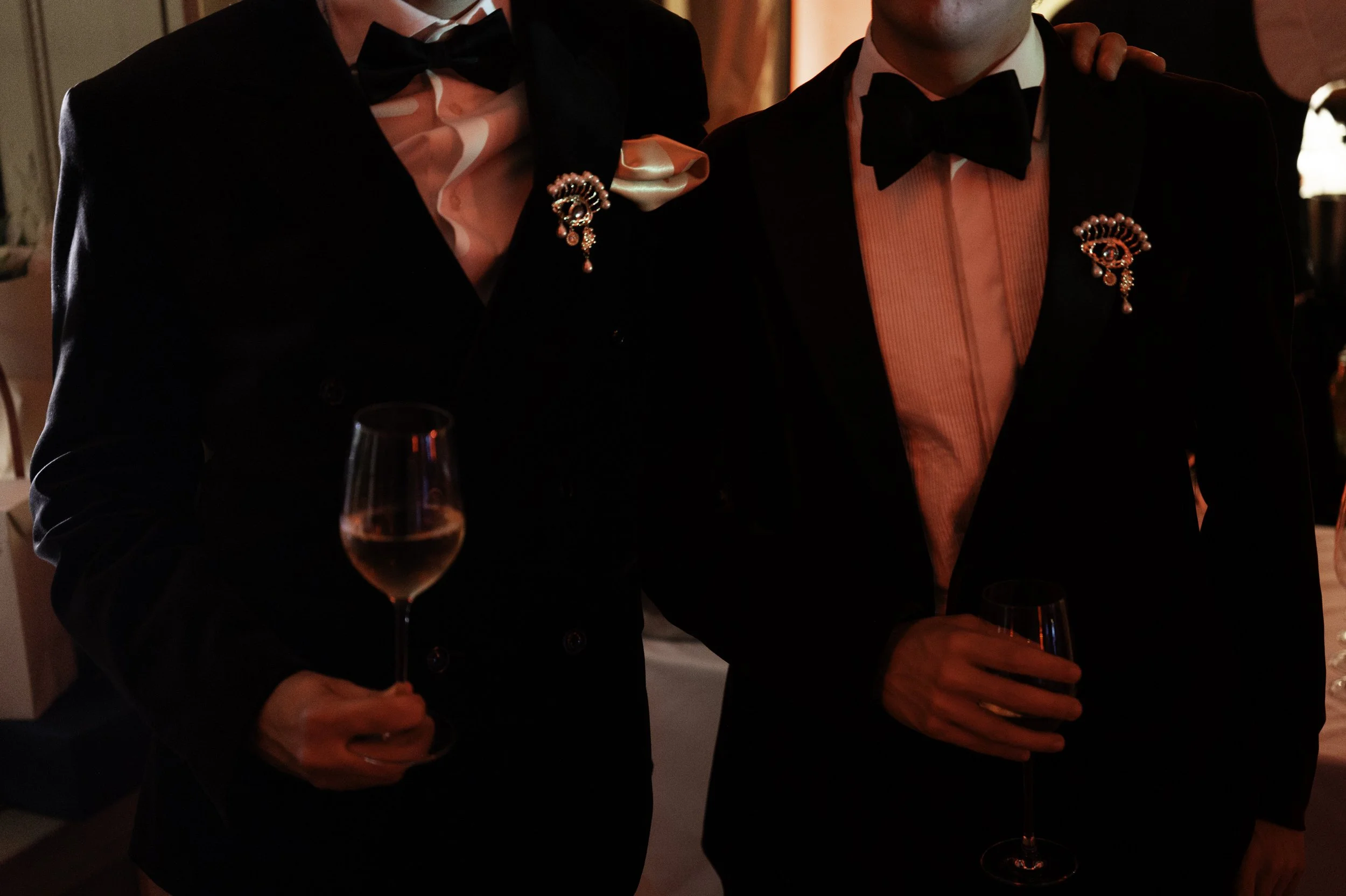 Two people dressed in tuxedos holding glasses of wine at a formal event. They have decorative brooches on their jackets and are standing in a dimly lit room with a red glow.