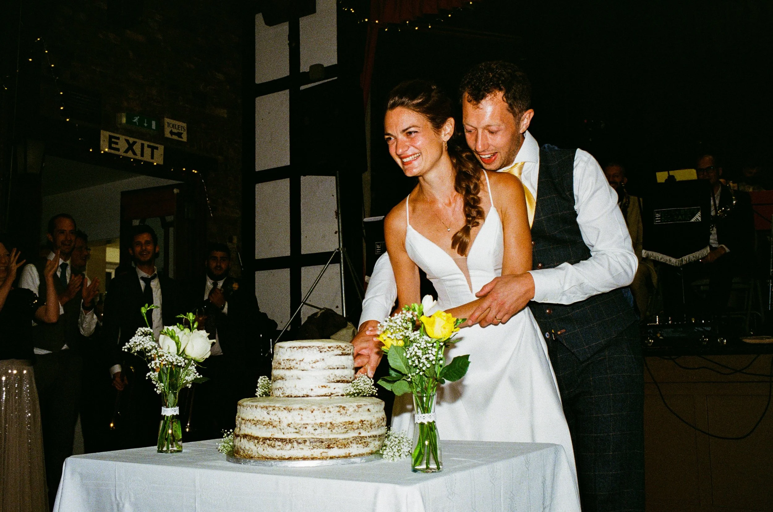 A couple is cutting a four-tier naked wedding cake decorated with white flowers and yellow roses, surrounded by small glass vases with white flowers on a white table. The bride is wearing a white dress and the groom is in a dark suit with a yellow ti