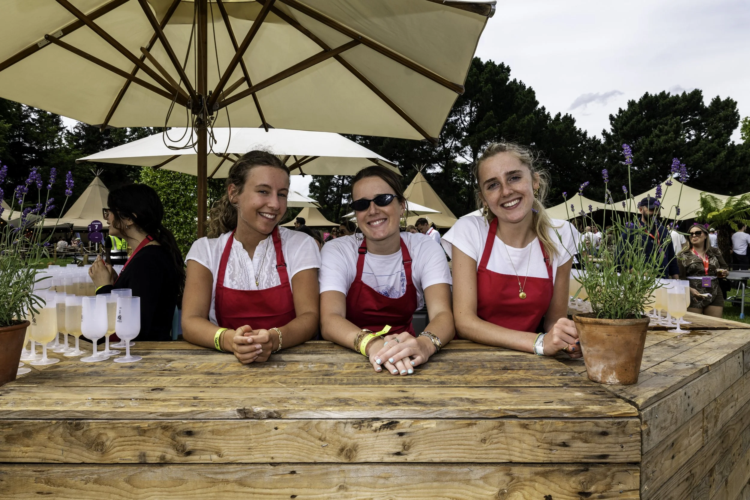 Three women smiling and sitting behind a wooden table at an outdoor event under large beige umbrellas, with plants and other people in the background.