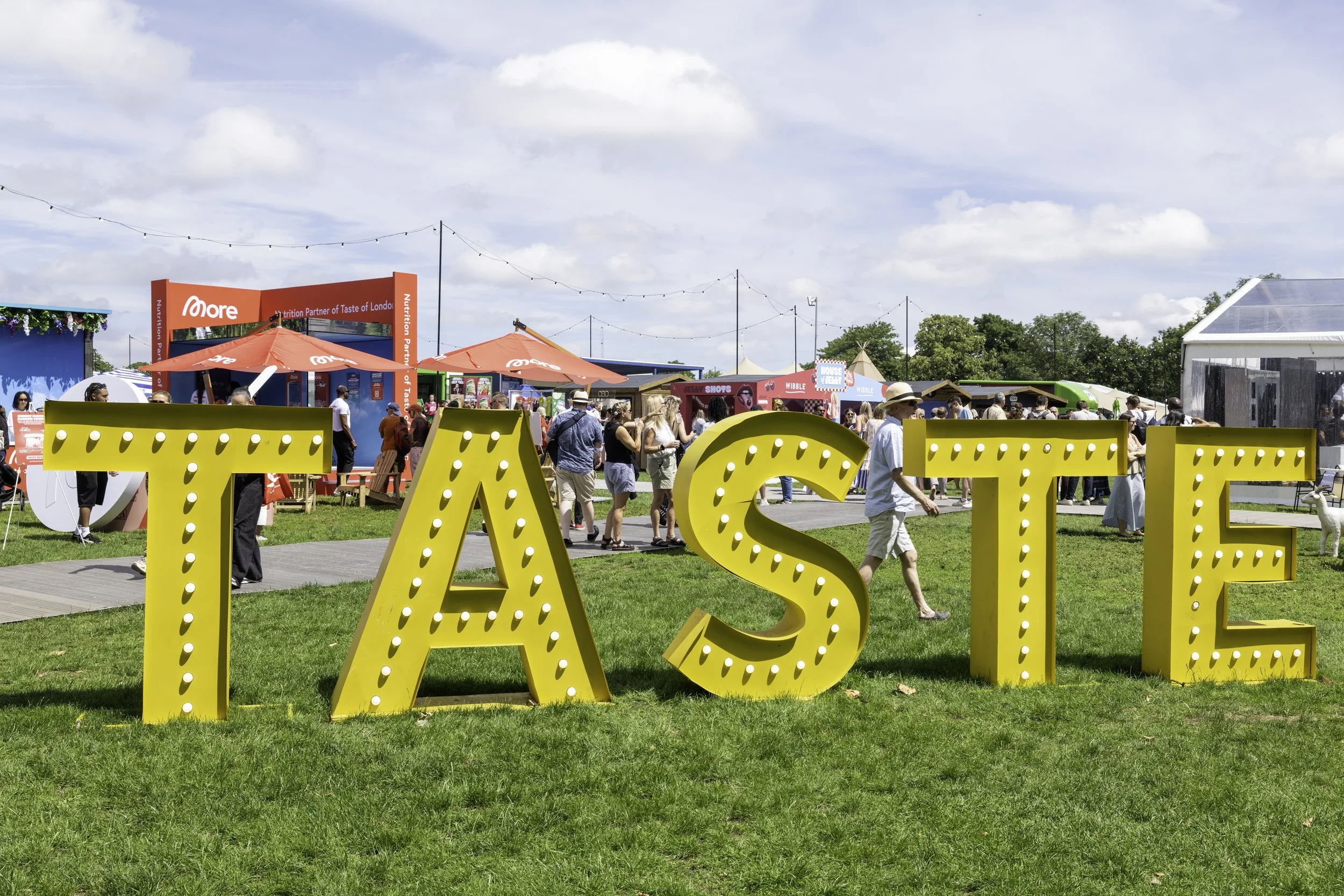 A lively outdoor event with people walking and gathering in a grassy area under a partly cloudy sky. Large yellow marquee letters spell out 'TASTE' in the foreground. Red umbrellas provide shade over vendor booths, and there are various colorful stal