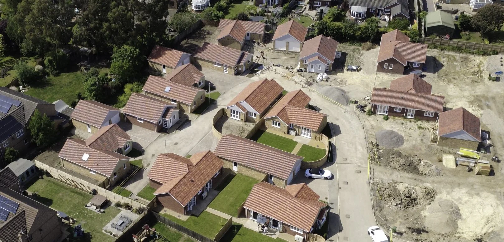 Aerial view of a residential neighborhood with several houses under construction, some finished with red-tiled roofs and others in various stages of building, surrounded by green lawns, trees, and a curved road.