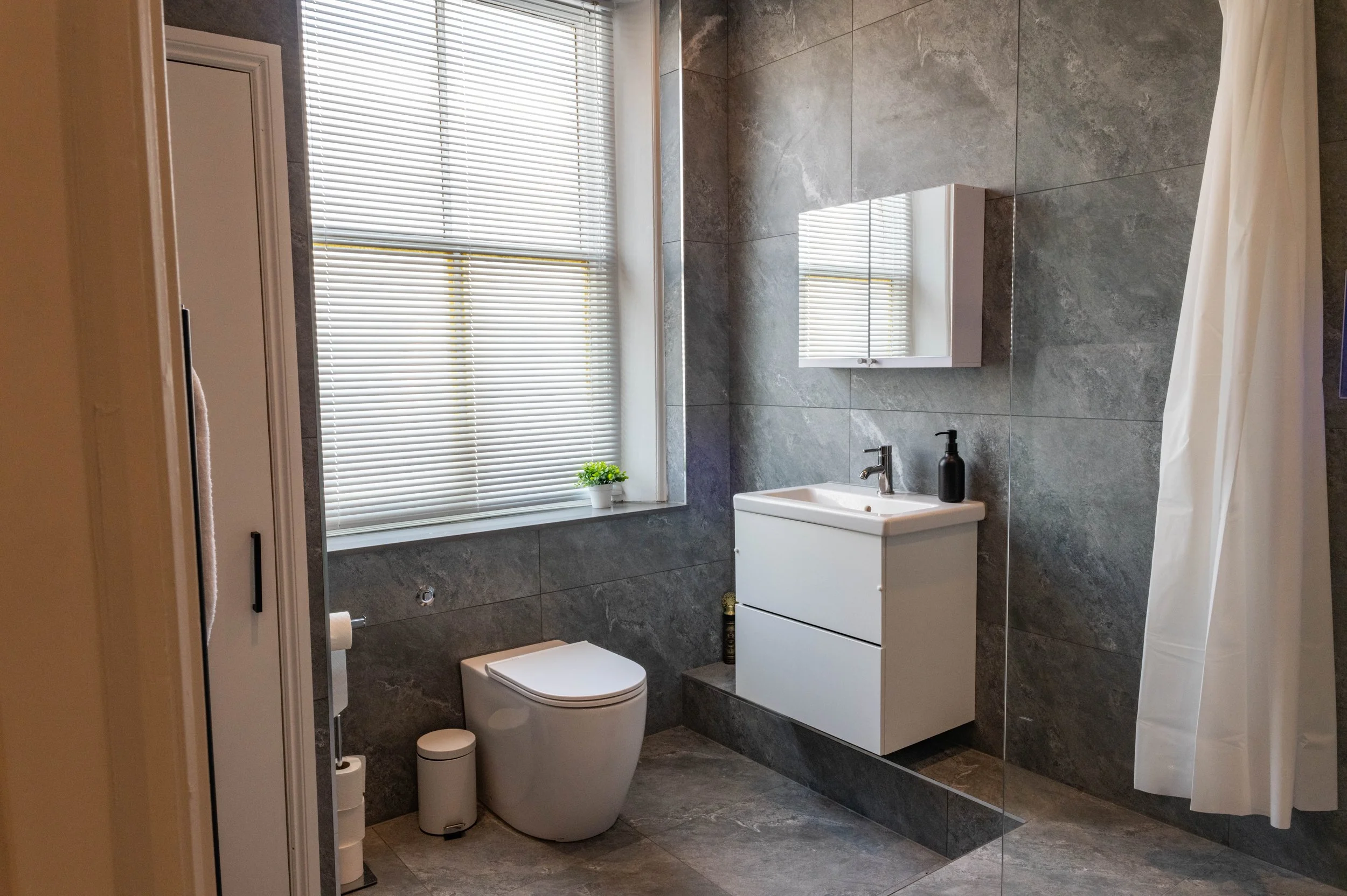 A modern bathroom with large gray tiles on the walls and floor. There is a window with white blinds, a small white cabinet with drawers, a sink with a black soap dispenser, and a white toilet. A small potted plant is on the window sill.
