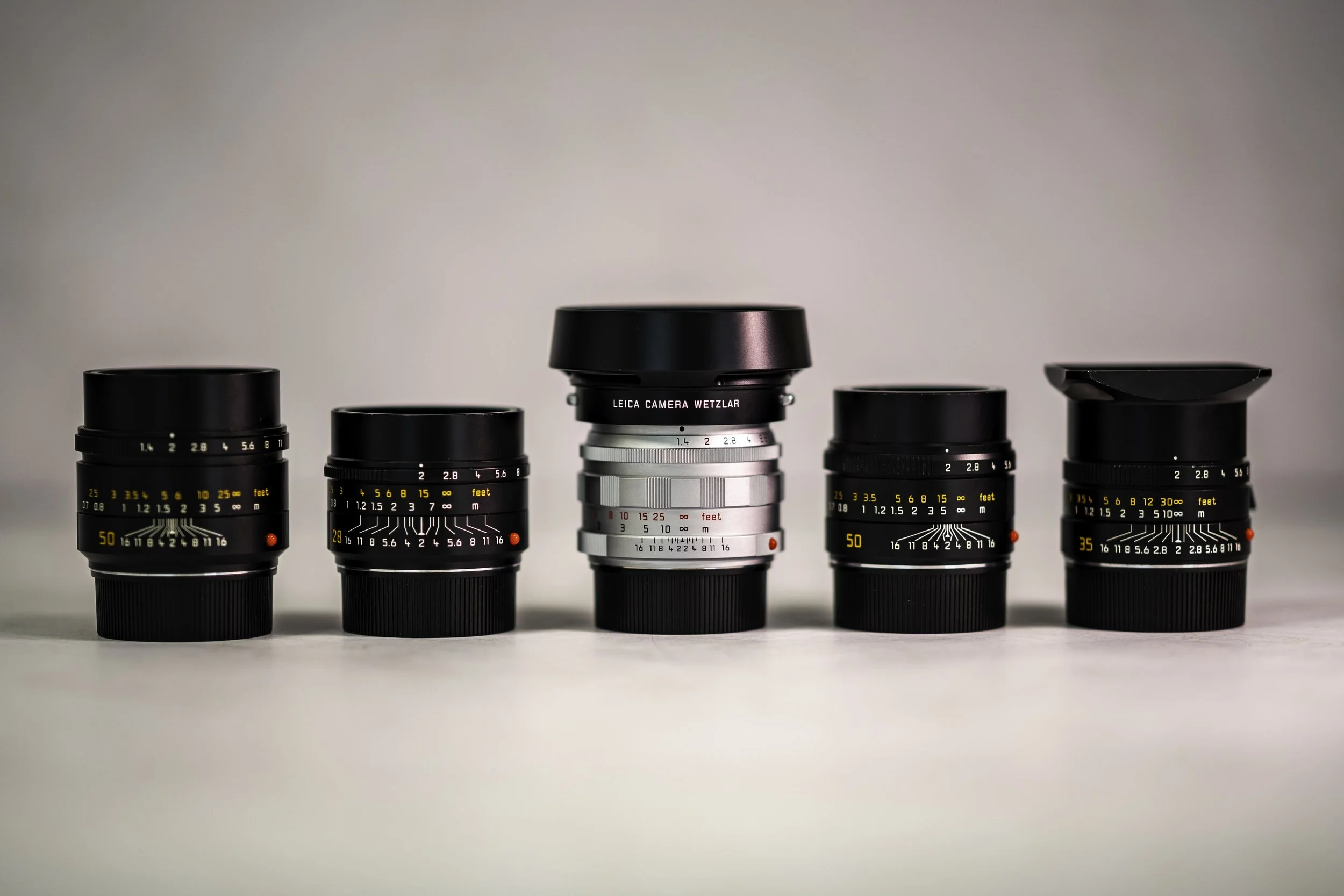 A lineup of five Leica  lenses on a neutral background.