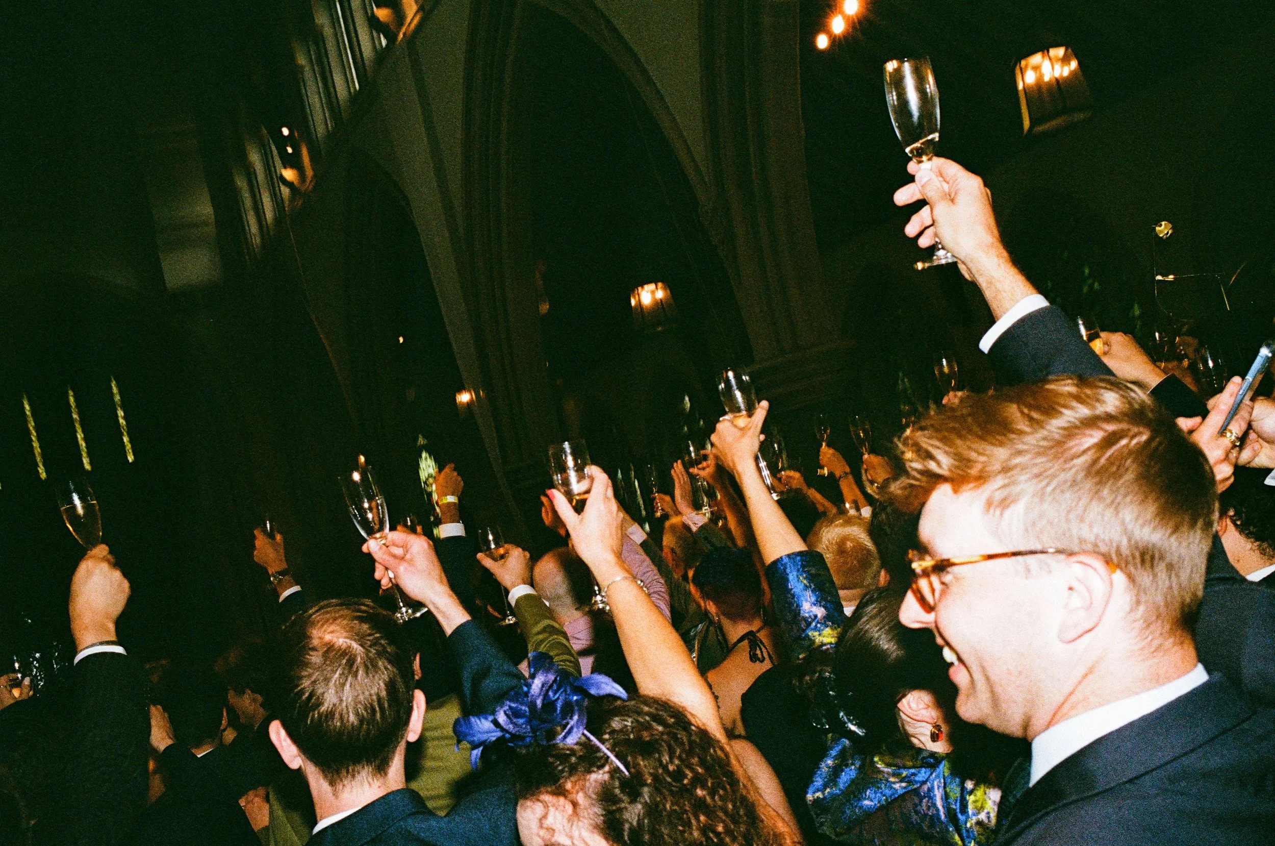 A group of people at a celebration raising champagne glasses in a toast, in an elegant indoor setting with warm lighting and high vaulted ceiling.