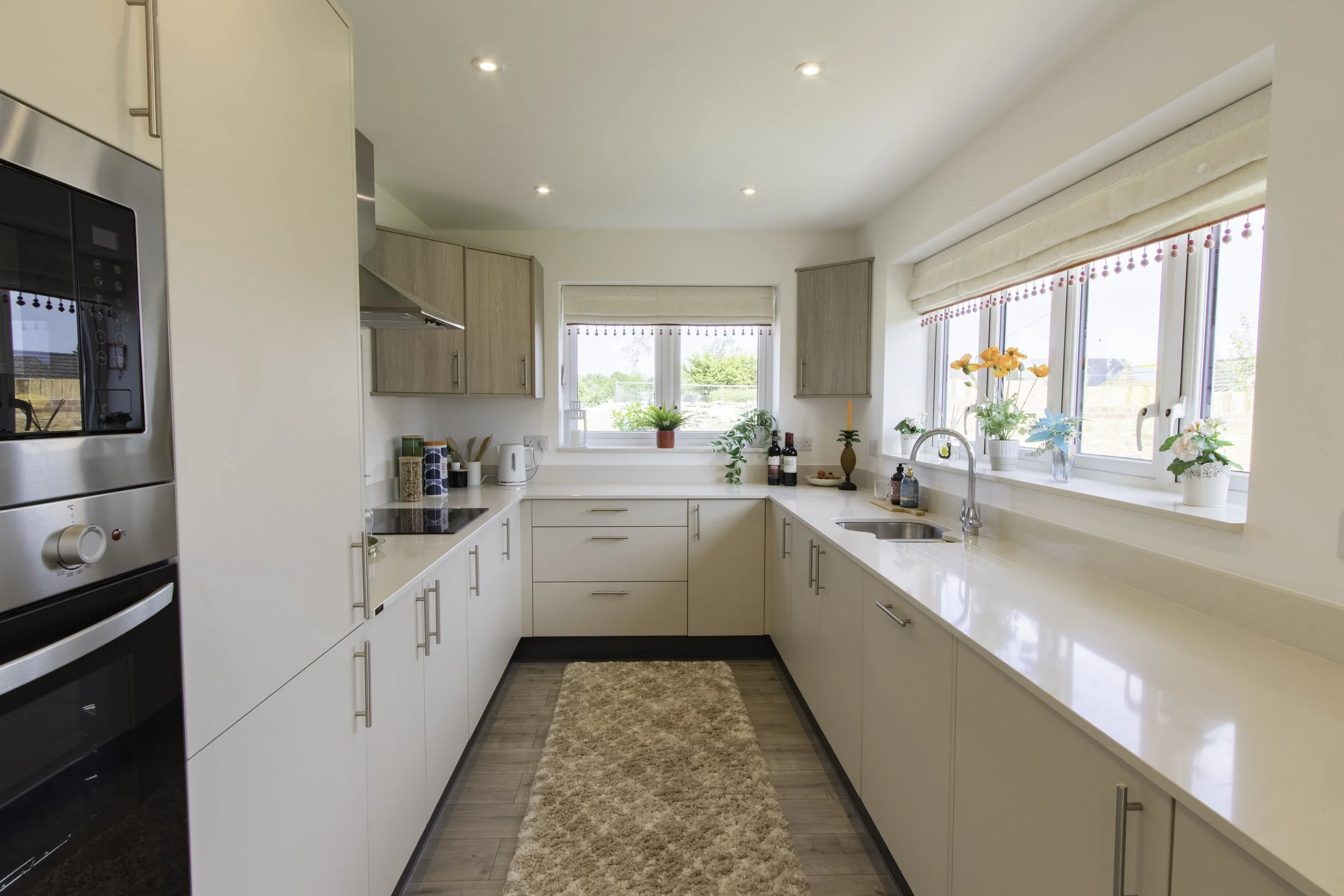 Bright kitchen with white cabinets, beige countertops, large windows with beige roman shades, and a wooden floor. Decor includes potted plants, flowers, and small kitchen appliances.