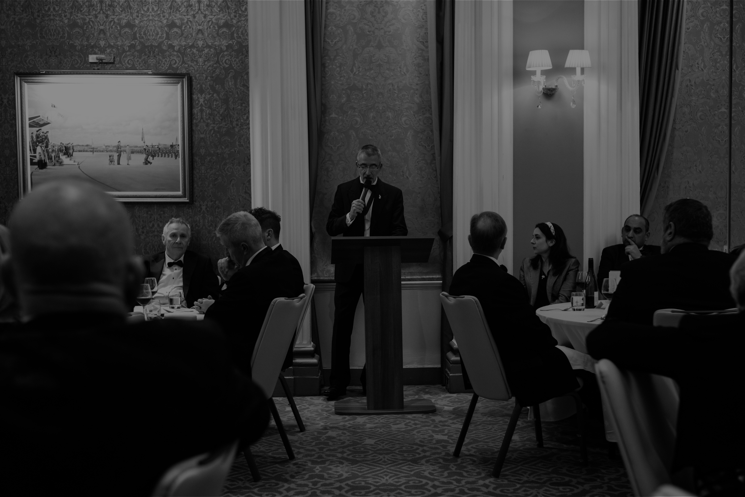 A  man in a tuxedo stands at a podium at aa black tie gala evening at a London gentleman's club, speaking into a microphone at a formal event, surrounded by seated guests in a decorated room with patterned wallpaper, a painting, and elegant lighting