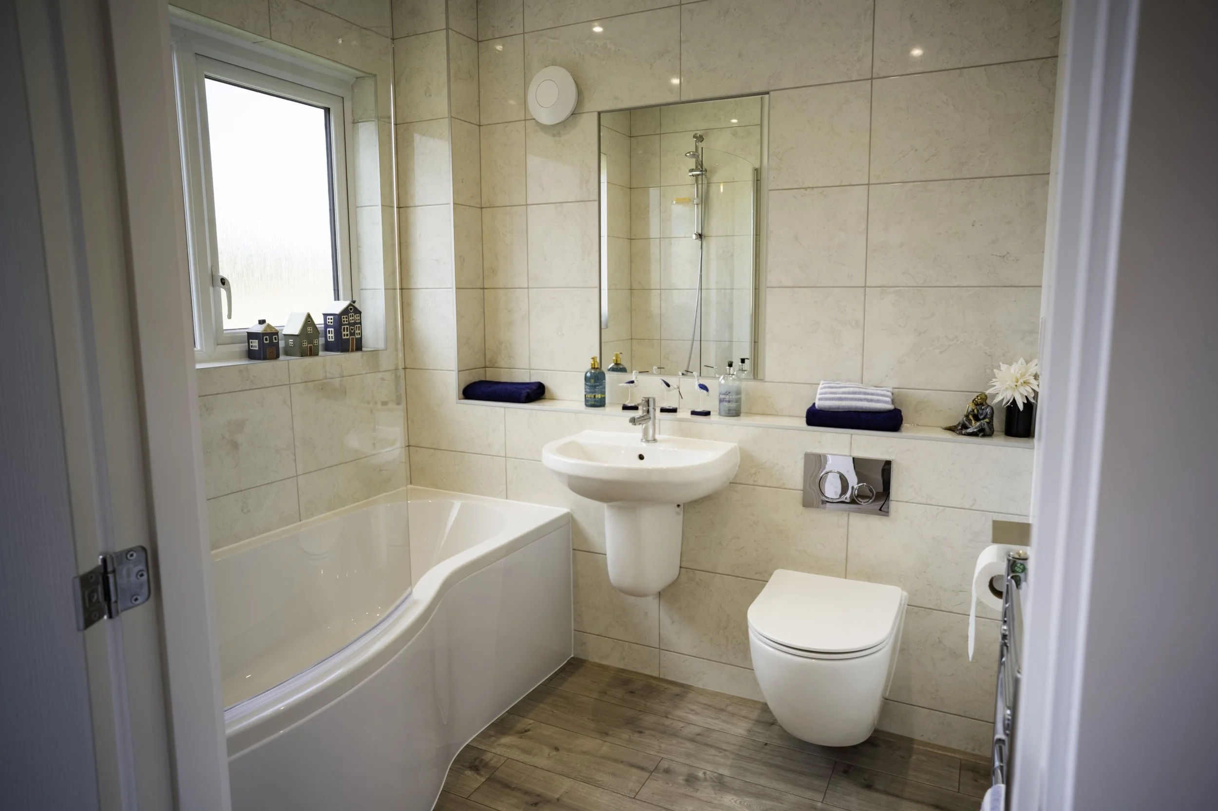 A clean modern bathroom with a bathtub, a toilet, and a wall-mounted mirror. The bathroom has beige tiled walls and a wooden floor. There are toiletries on the sink and a small decorative sculpture with a flower on the right side.