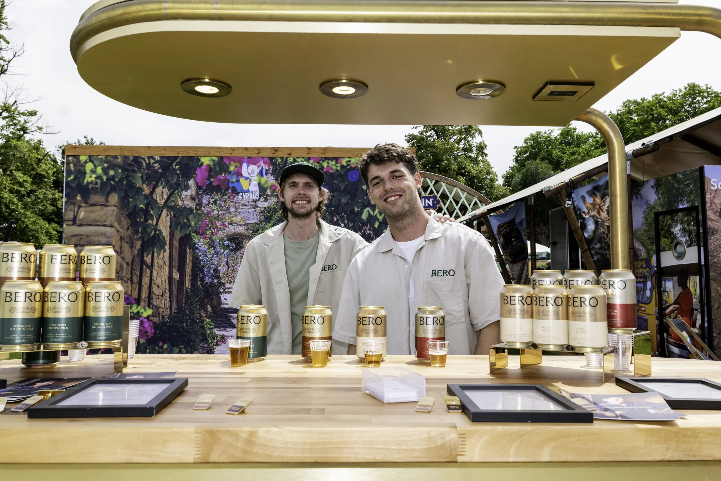 Two men standing behind a wooden table with cans of BERO beer, outdoors at a festival or event, with trees and stage backdrop behind them.