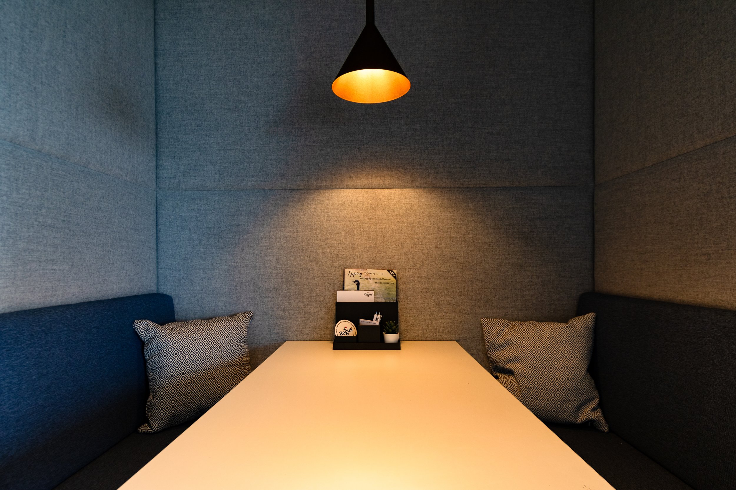 Interior of a cozy booth with blue fabric walls, a white table, patterned cushions, and a black and orange pendant light overhead.