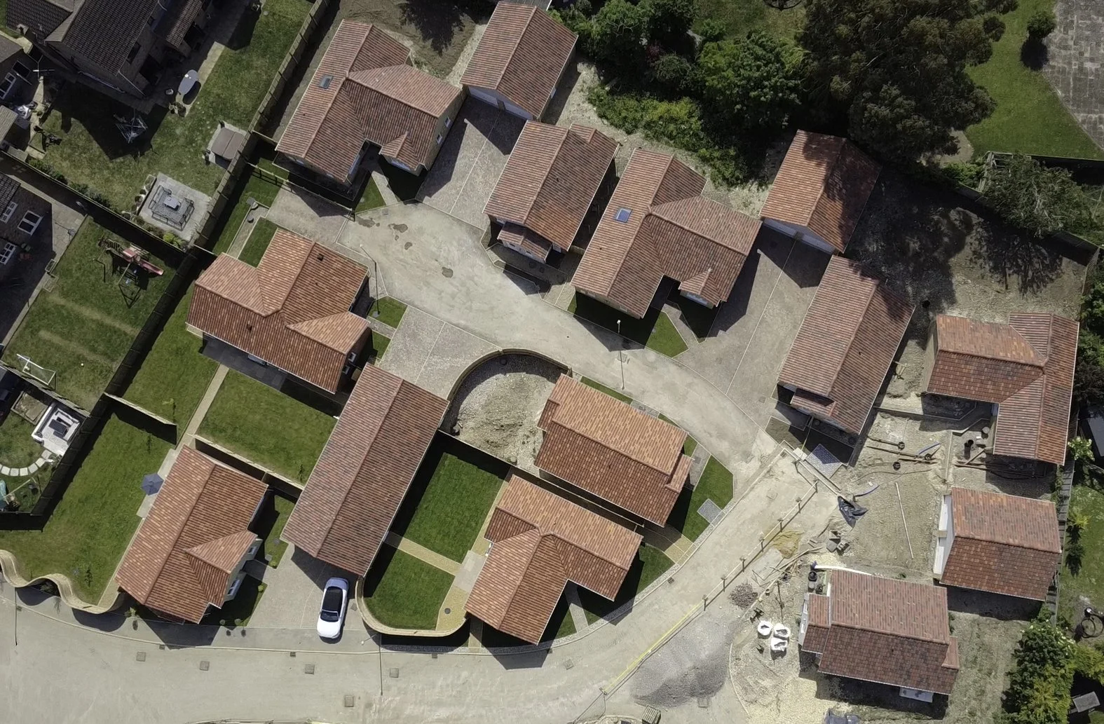 An aerial view of a residential complex with multiple houses featuring red-tiled roofs, green lawns, a parking area, and some ongoing construction.