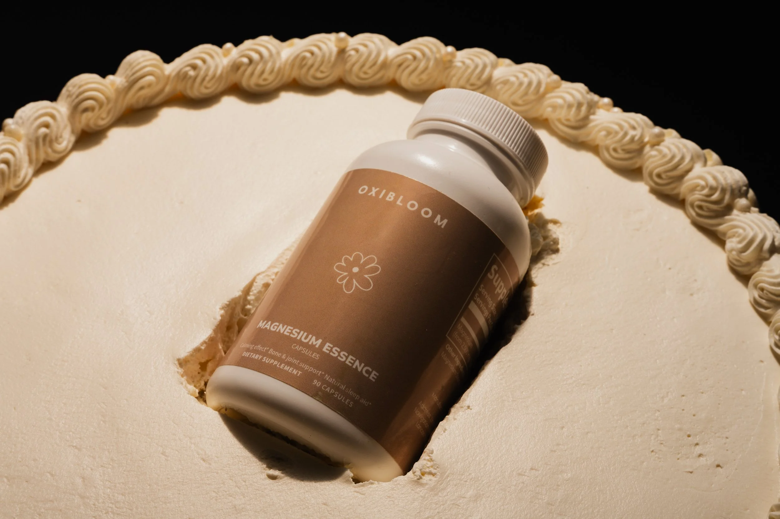A bottle of OXIBLOOM magnesium essence supplement resting on a white cake with a decorative border of piped cream, half of a hole cut into the cake.