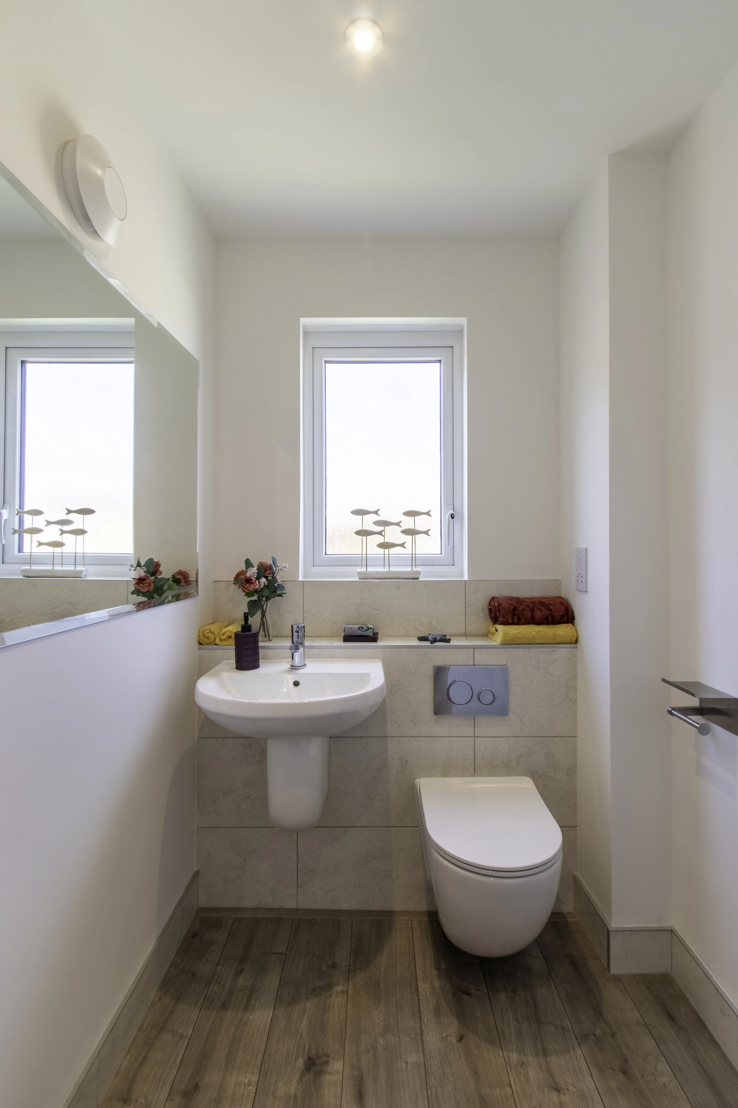 A modern bathroom with a white sink, toilet, and a window with natural light. Decor includes flowers, towels, and small decorative plants. Wooden flooring and minimalistic fixtures.