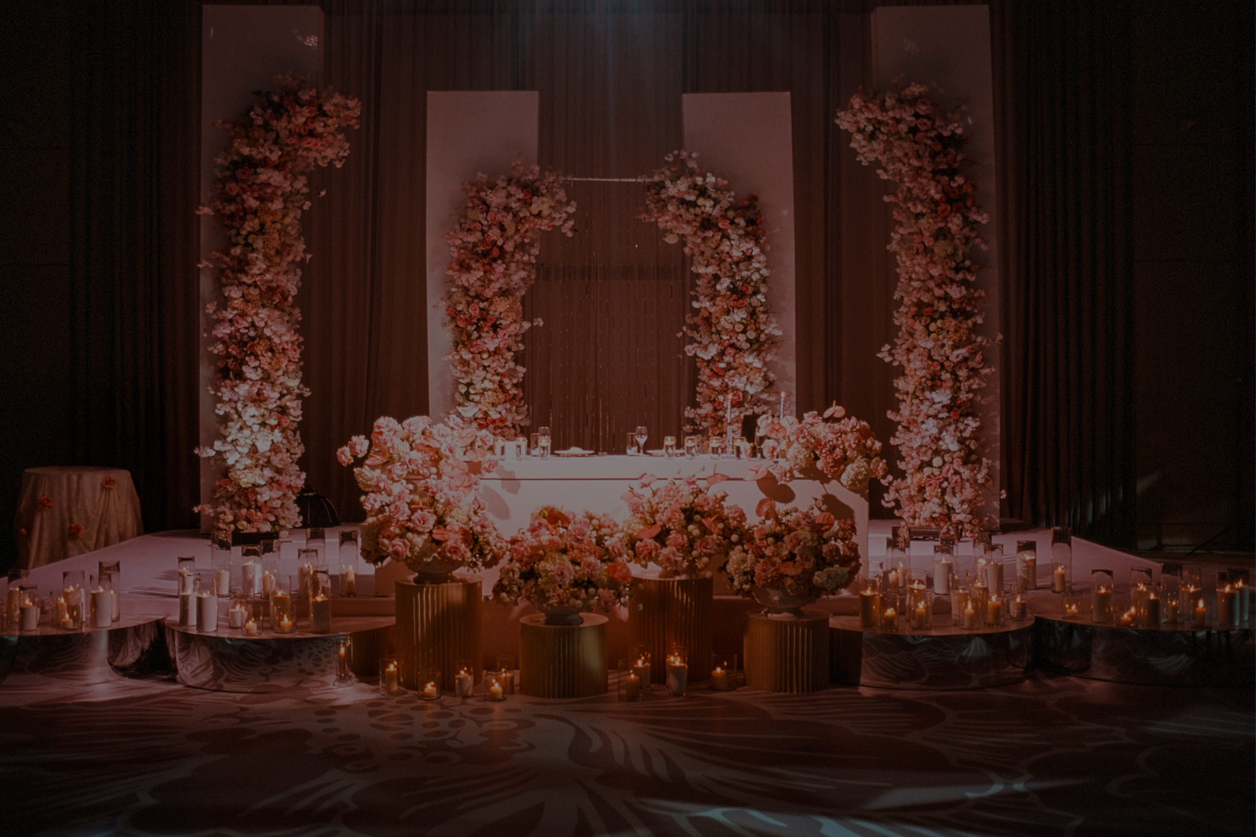 Indoor luxury floral wedding or event decor with pink and white floral arrangements, candles, and draped curtains, stage with a floral arch and table.