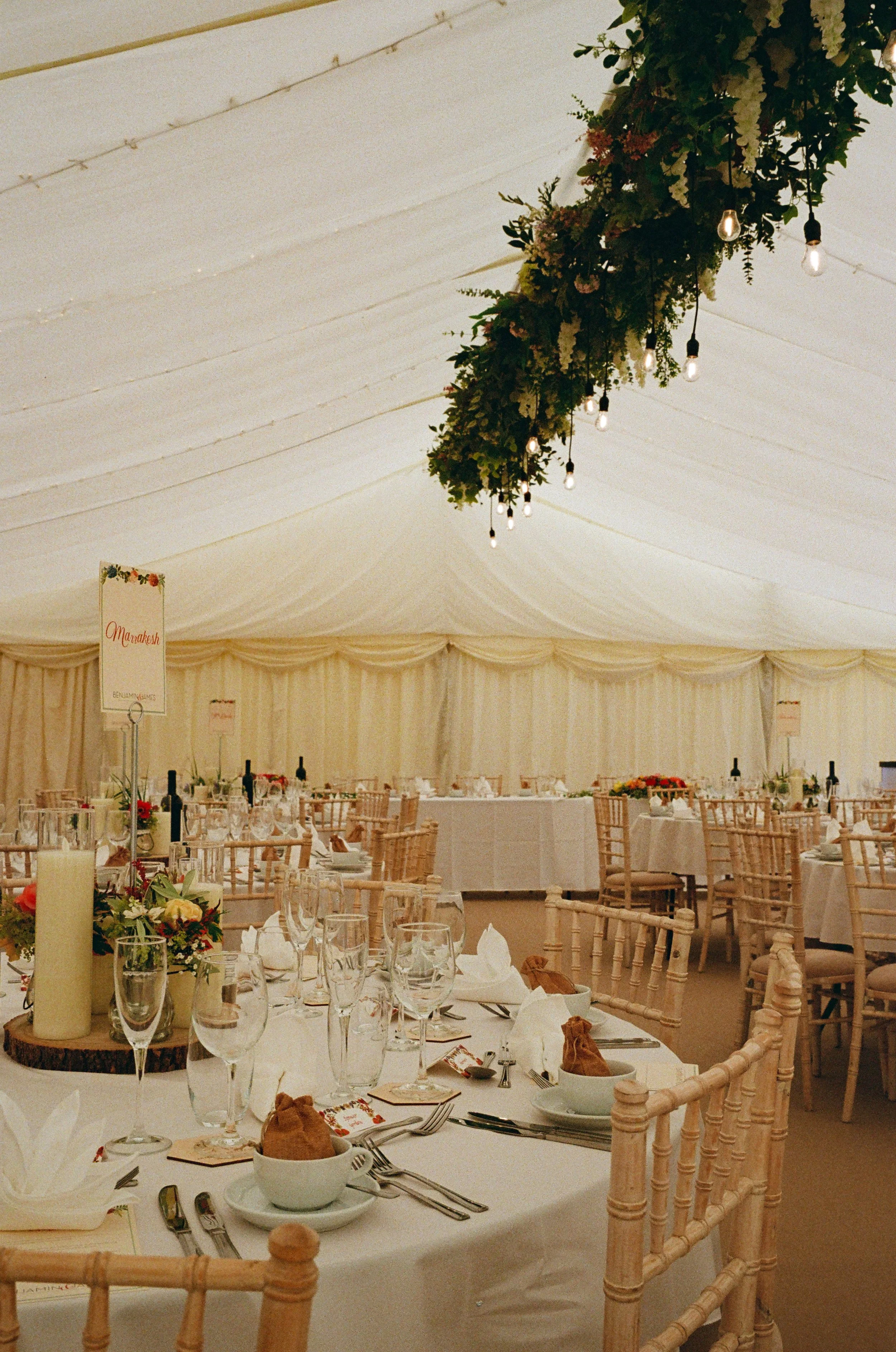 Interior of a wedding reception tent with decorated tables, floral centerpieces, and hanging lights.