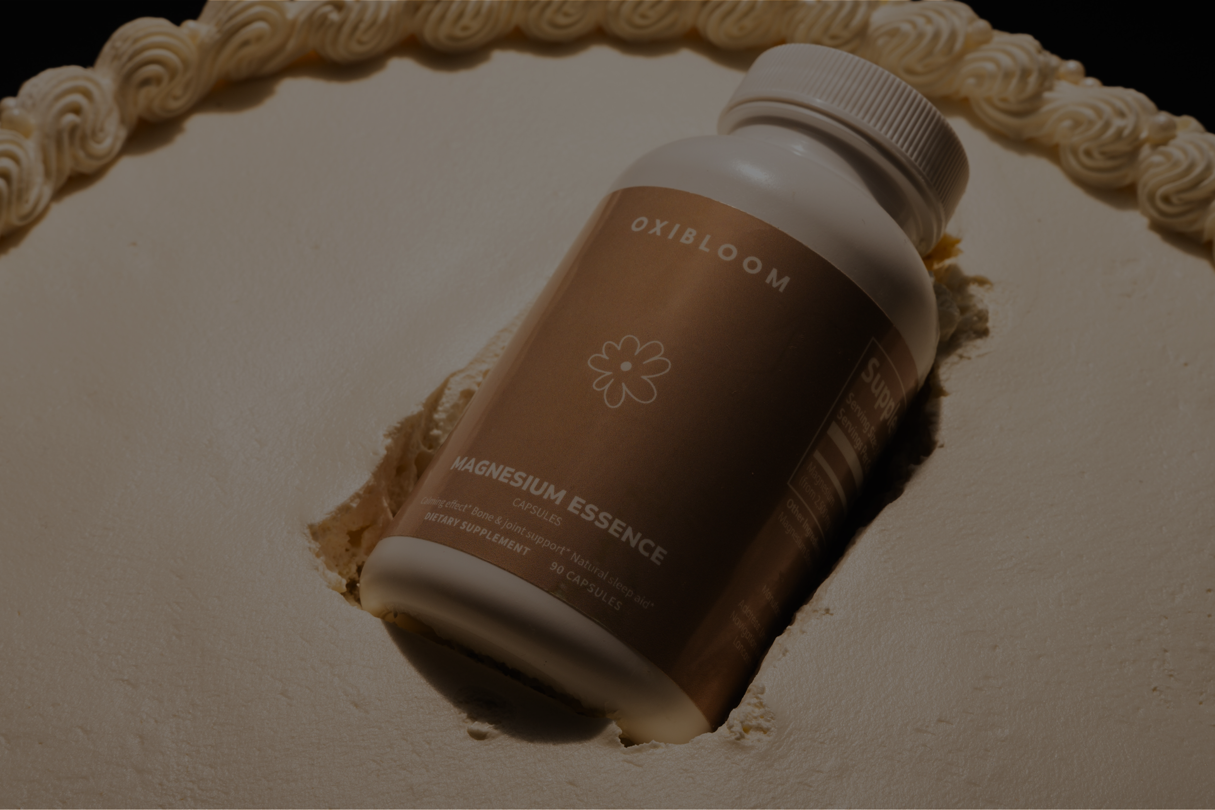 A premium product photography of a bottle of magnesium essence capsules lying in a hole in a white cake decorated with cream swirls around the edge.
