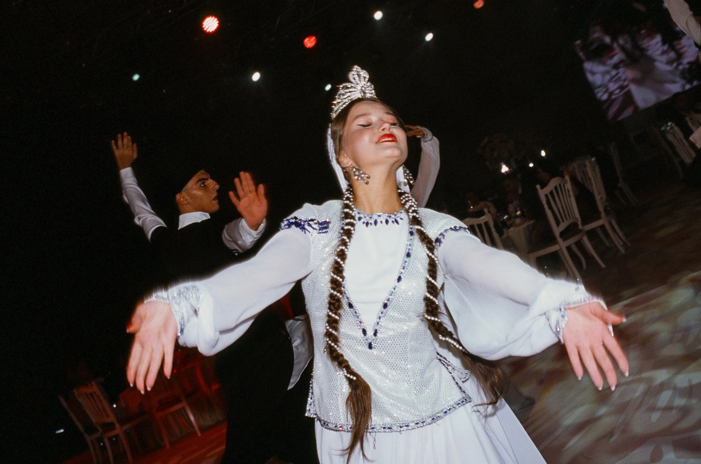 A woman wearing traditional attire and a crown is dancing with her eyes closed, others in formal clothing are visible in the background.