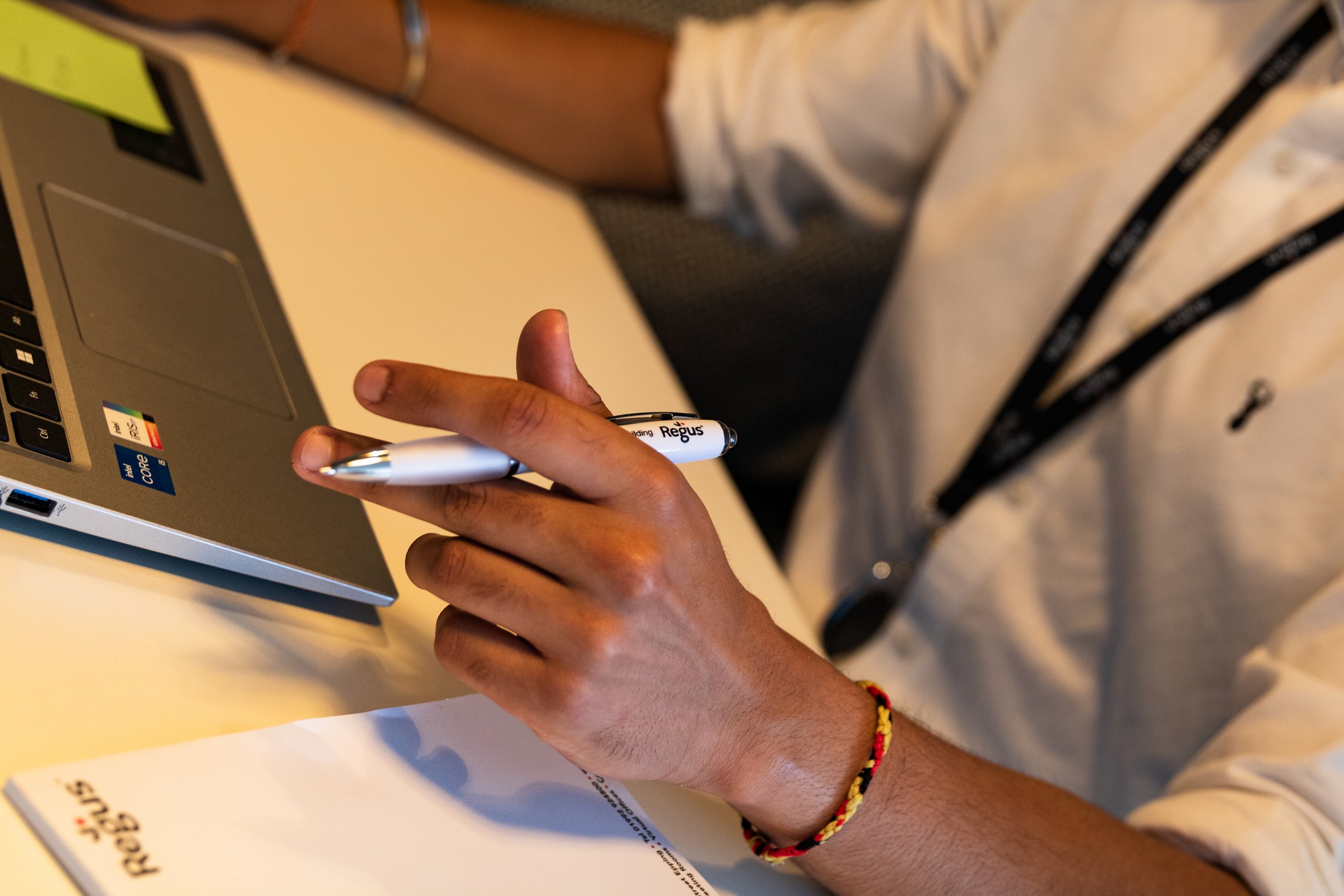 A person holding a white pen with black and red text that says 'Regus' near a silver laptop with an Intel Core i5 sticker. The person's arm has a colorful braided bracelet. The background is a blurred indoor setting.