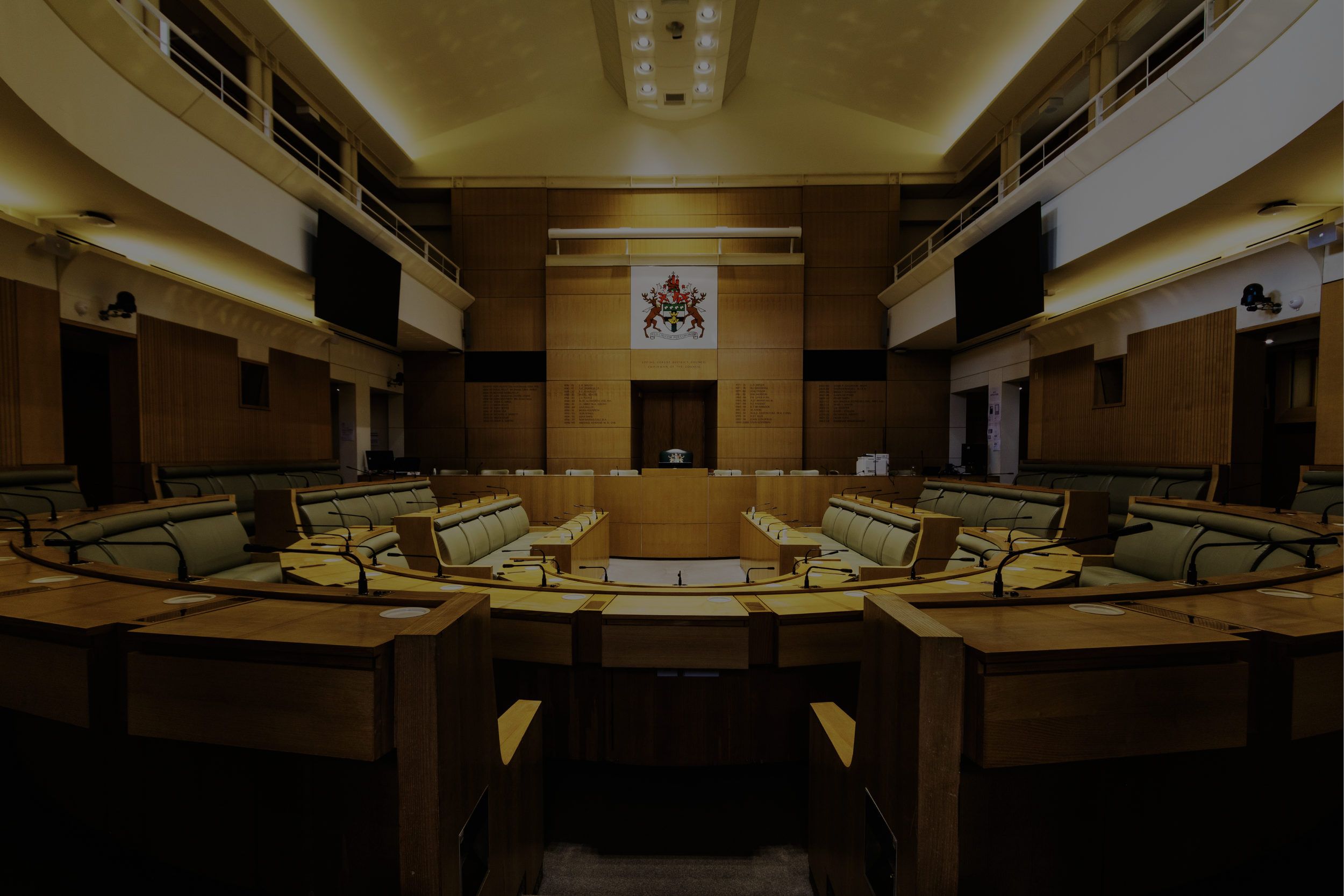 A large, empty conference or legislative chamber with a semi-circular seating arrangement, wood paneling, multiple large screens, and a crest or emblem on the front wall.