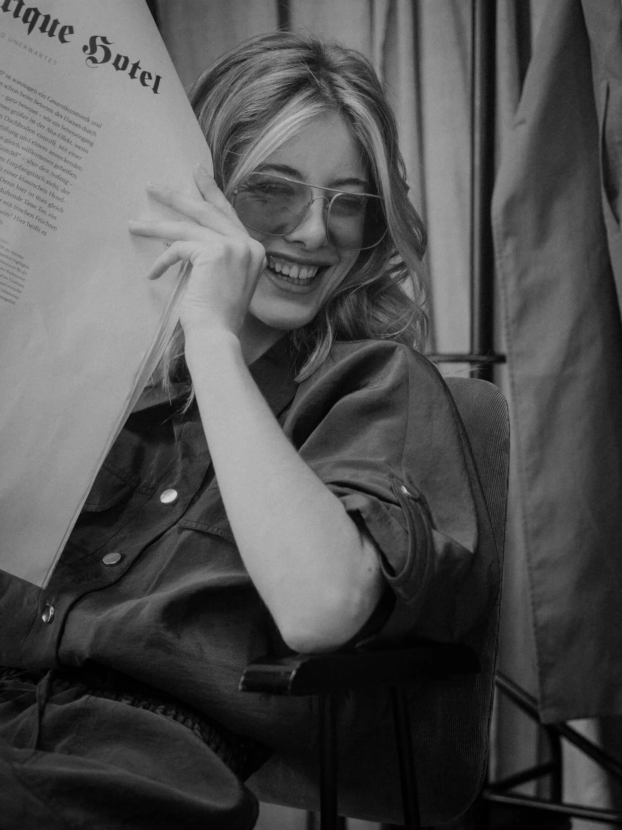 Junge Frau mit Sonnenbrille, lächelnd, hält einen Zeitungspapier vor Gesicht, sitzend auf einem Stuhl, im Hintergrund Vorhänge.