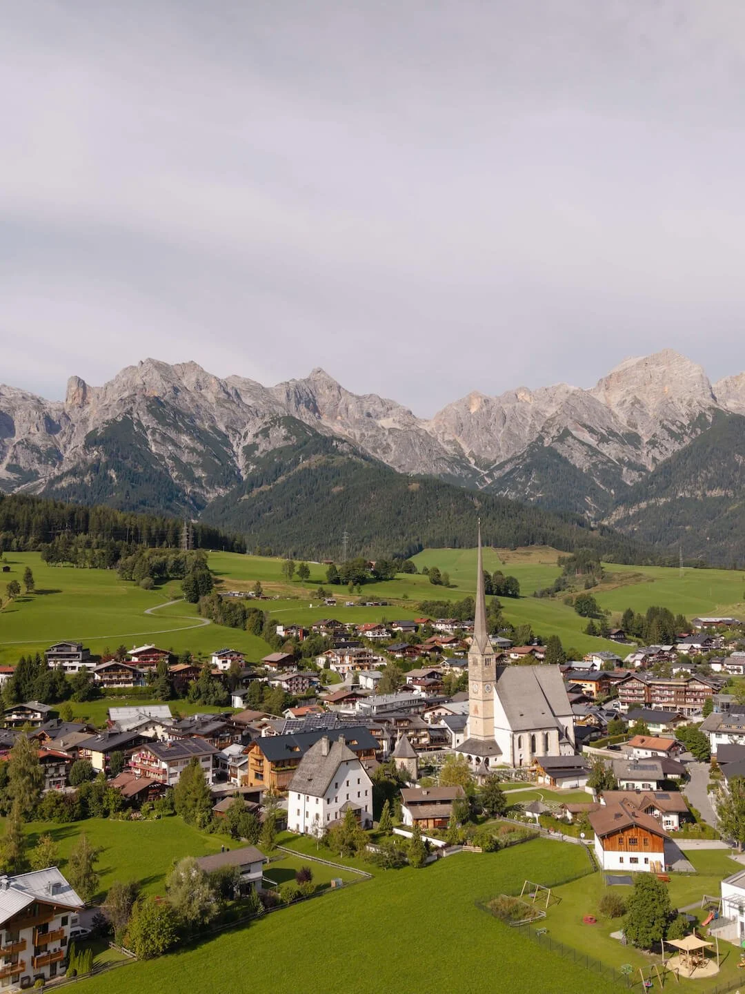 Das Dorf Maria Alm am Hochkönig in einer bergigen Landschaft mit einer Kirche im Zentrum, umgeben von grünen Feldern und Häusern, mit hohen Bergen im Hintergrund.