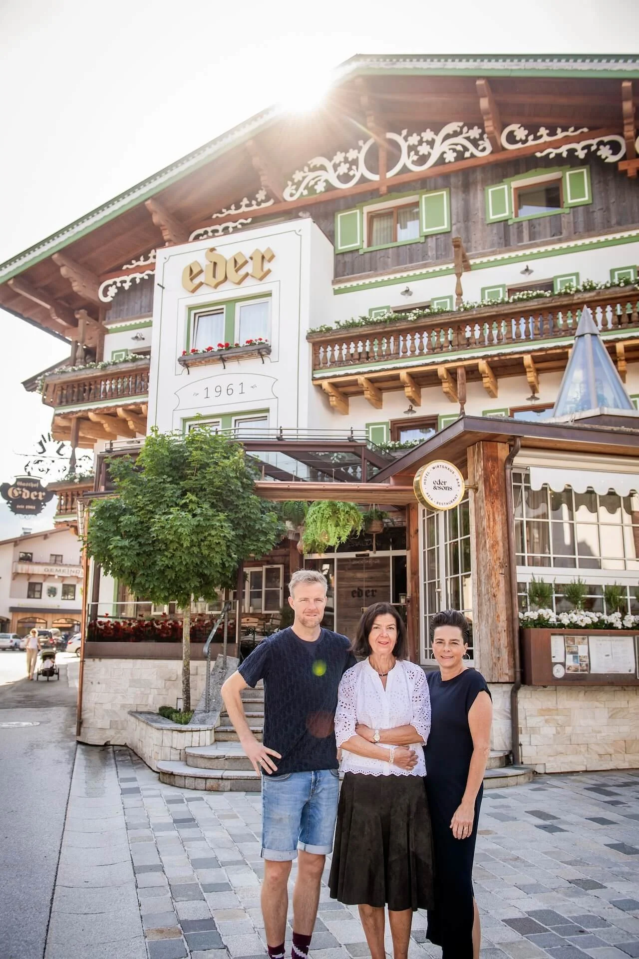 Drei Menschen stehen vor einem traditionellen bayerischen Gasthaus namens "Eder" mit Holzelementen und Blumenkästen auf den Fenstern. Es ist ein sonniger Tag, und die Gruppe posiert für das Foto.