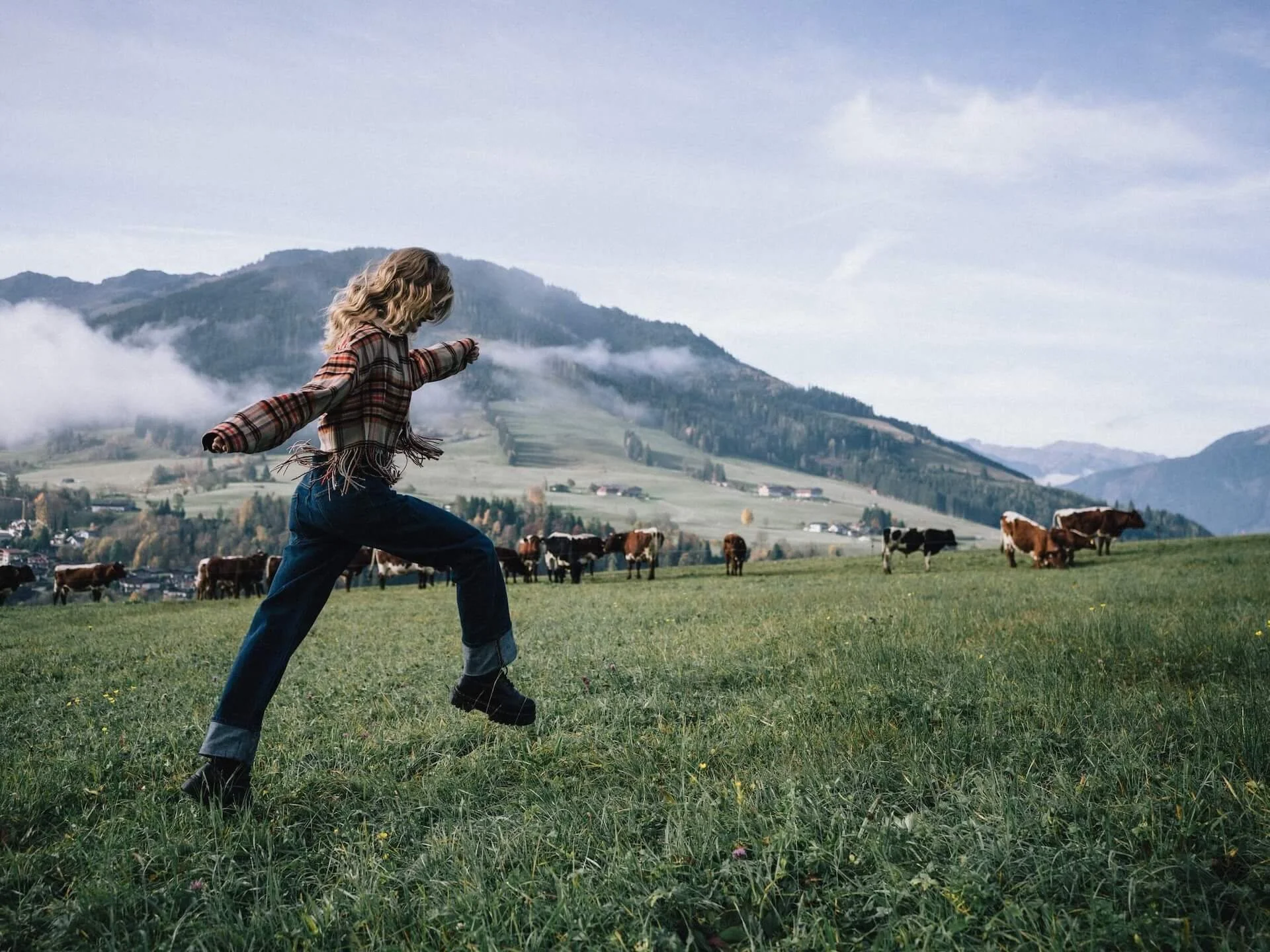 Ein Kind in Jeans, Barfuß und mit kariertem Hemd spielt auf einer grünen Wiese mit Kühen vor einer Berglandschaft, Wolken und Himmel im Hintergrund.
