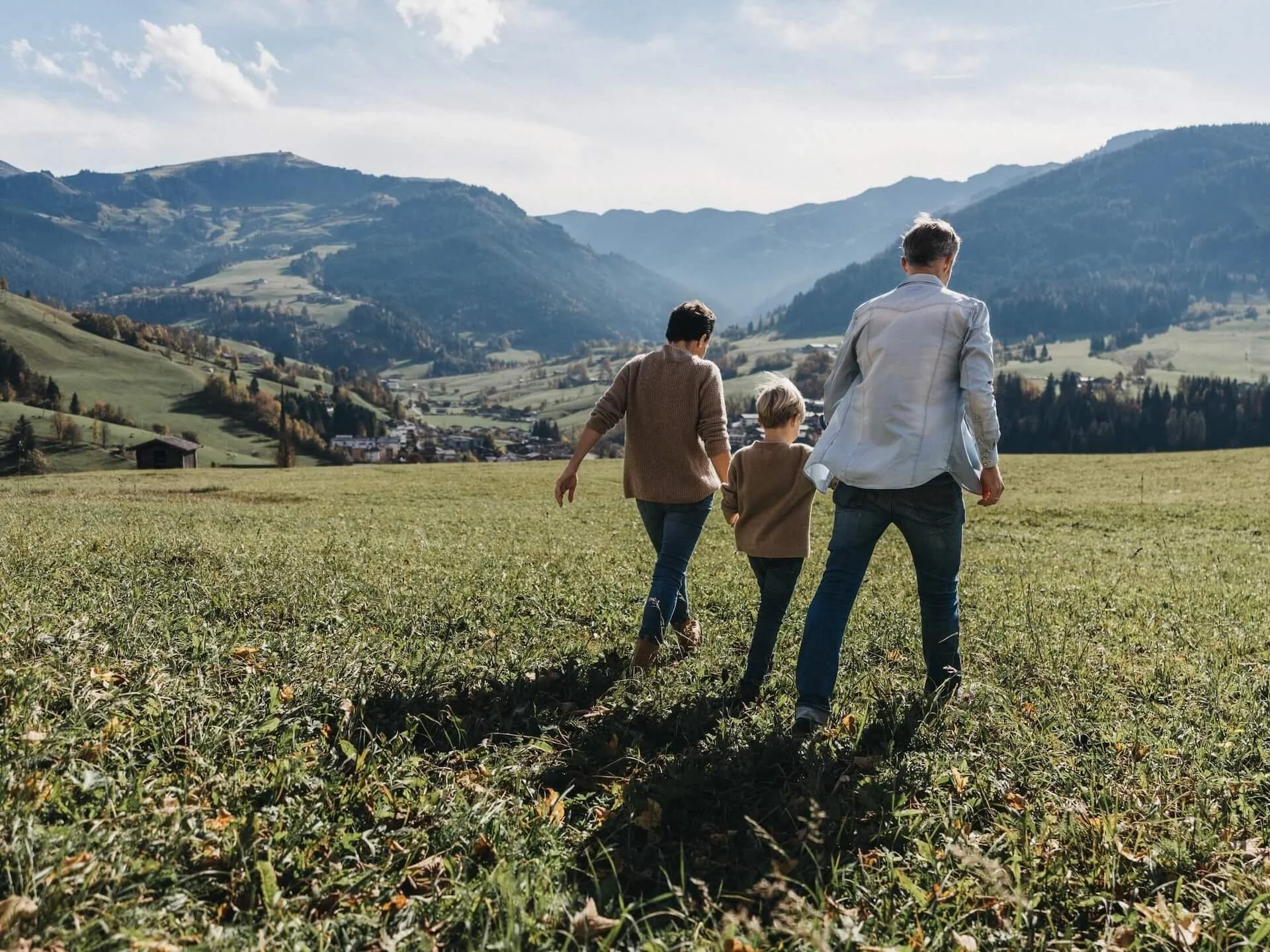 Drei Personen gehen durch ein grünes Feld in den Bergen, mit einem kleinen Dorf im Hintergrund.