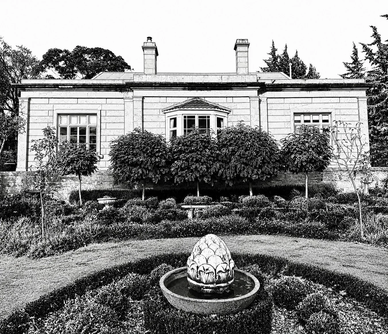 Black and white photo of a house with a garden in front, featuring a fountain with a decorative stone sculpture in the center of a circular flower bed.