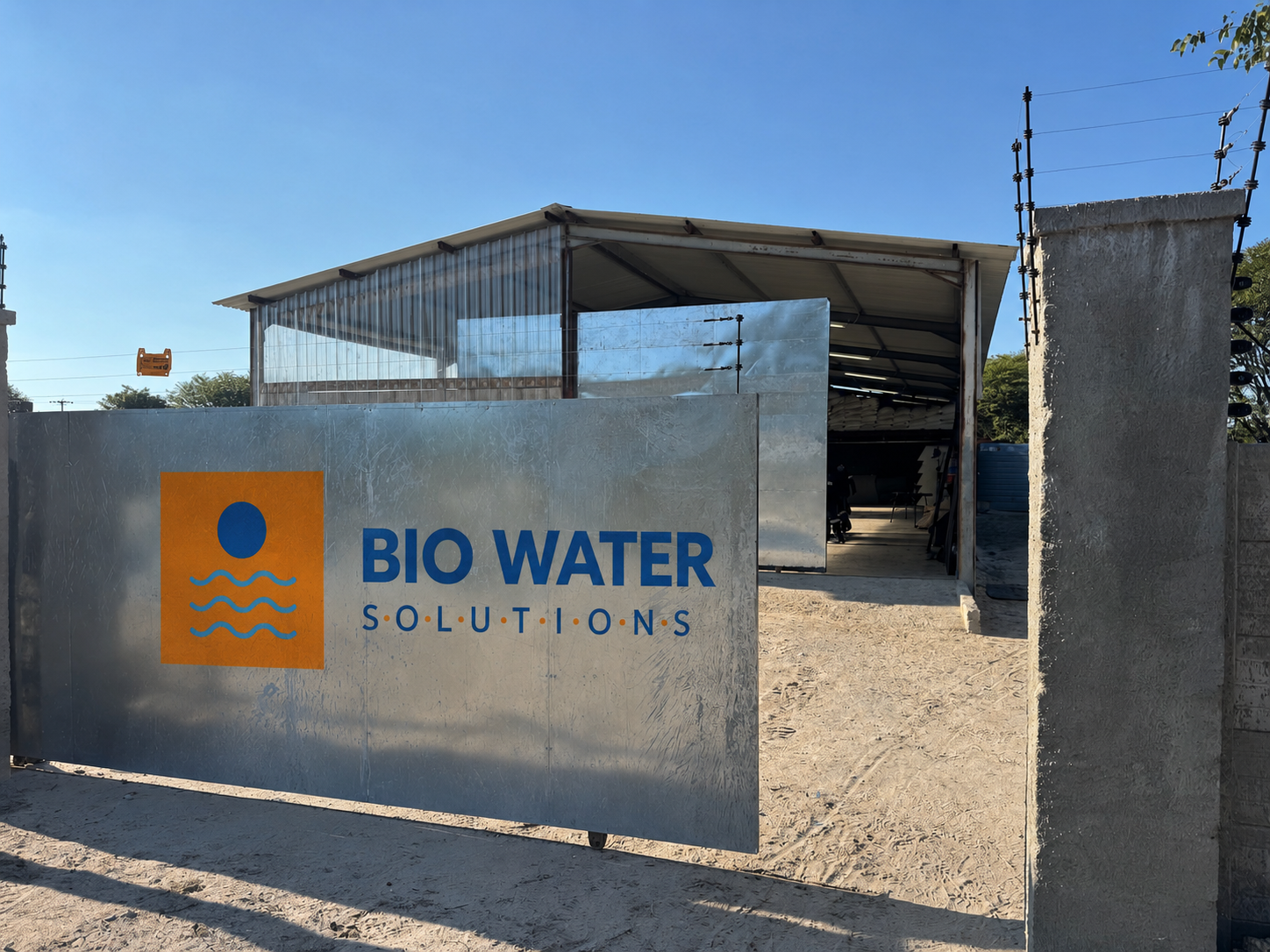 Entrance to Bio Water Solutions facility with a sign on a metal gate, showing a sun and water waves logo, and a large open-sided building inside.