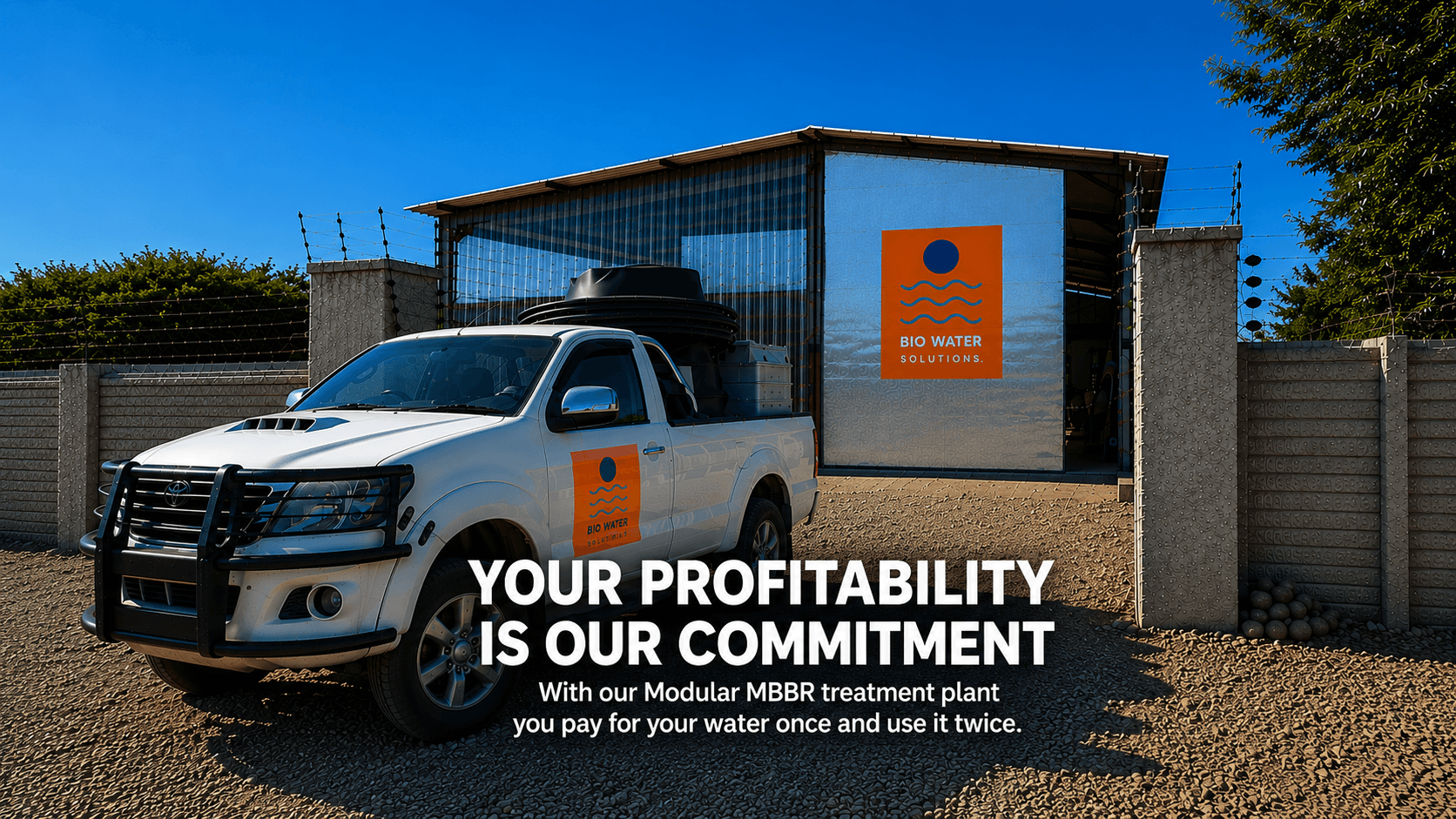 A white truck with a bio water solutions logo on the door parked in front of a modular treatment plant with another logo on the building, under a clear blue sky, with a dirt ground and a fence with barbed wire.
