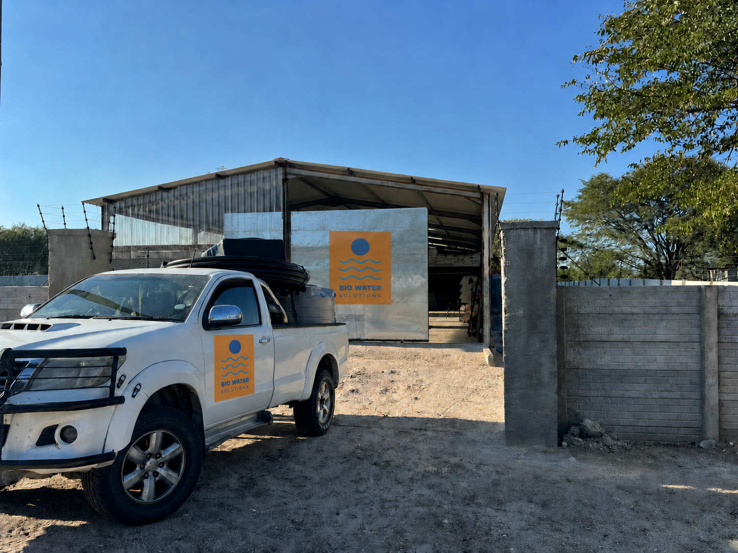 A white pickup truck with 'Bio Water Solutions' logo on the door parked outside a semi-open workshop with the same logo on a sign on the building. The area is sandy with some greenery and a concrete wall with barbed wire on top.