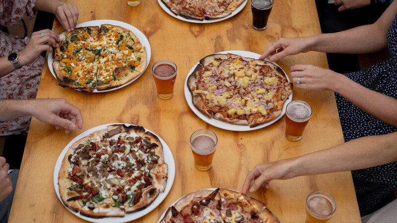 Multiple pizzas and glasses of beer on a wooden table with people reaching for the pizza slices.