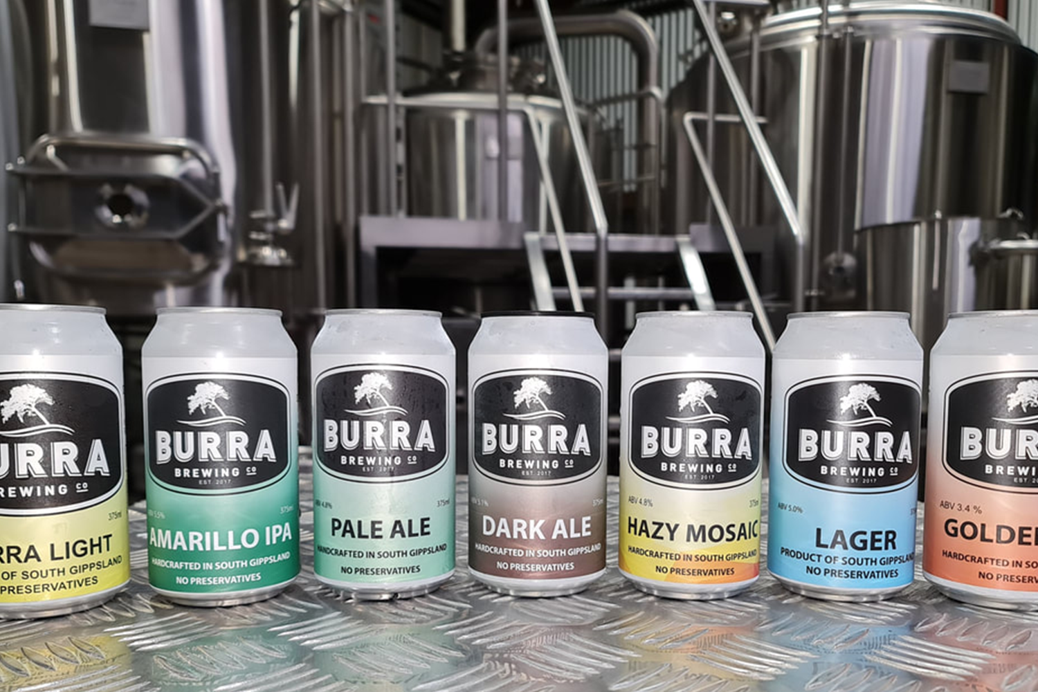 Six cans of beer from Burra Brewing Co. on a metal surface in front of brewing equipment; flavors include Light, Amarillo IPA, Pale Ale, Dark Ale, Hazy Mosaic, Lager, and Golden, each labeled with alcohol content and brewed in South Gippsland.