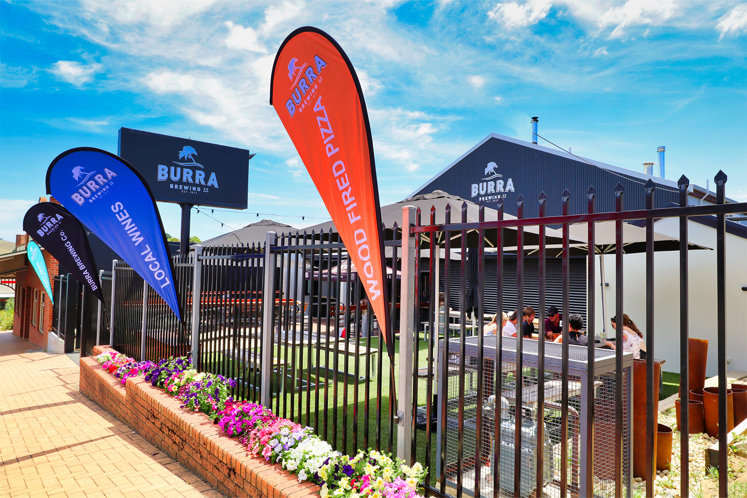 Outdoor patio of Burra Brewing Co with banners advertising local wines, wood-fired pizza, and the brewery name, surrounded by colorful flowers and a black metal fence, with people sitting at tables inside.