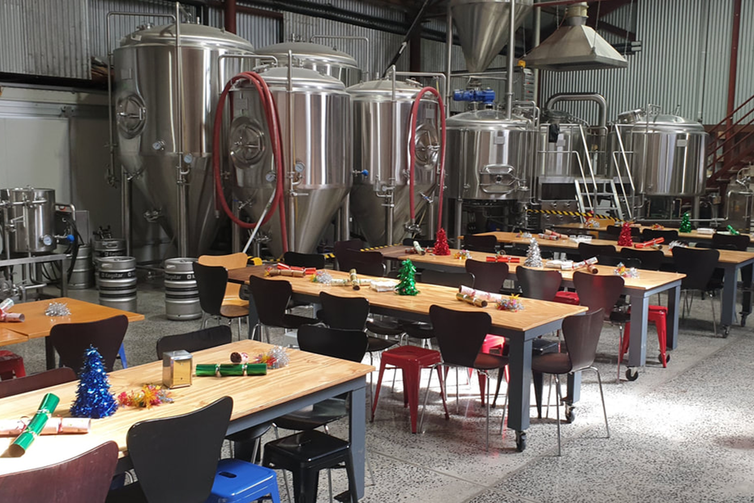 Inside a brewery decorated for Christmas with tables, chairs, and holiday decorations such as small Christmas trees, ornaments, tinsel, and party blowers.