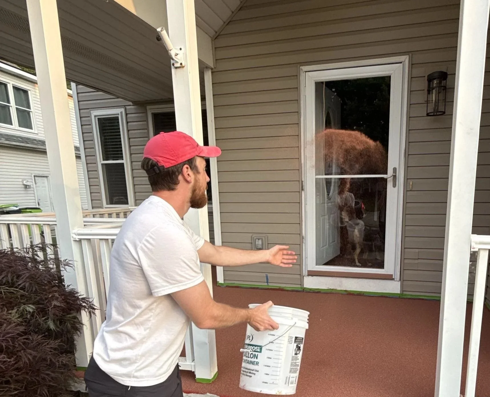 A man is outside on a porch, tossing a bag of cat litter through a screen door toward a cat standing inside. The man is wearing a white t-shirt and a pink baseball cap, and is holding a white container. The porch has outdoor furniture and the house's exterior is beige with siding.