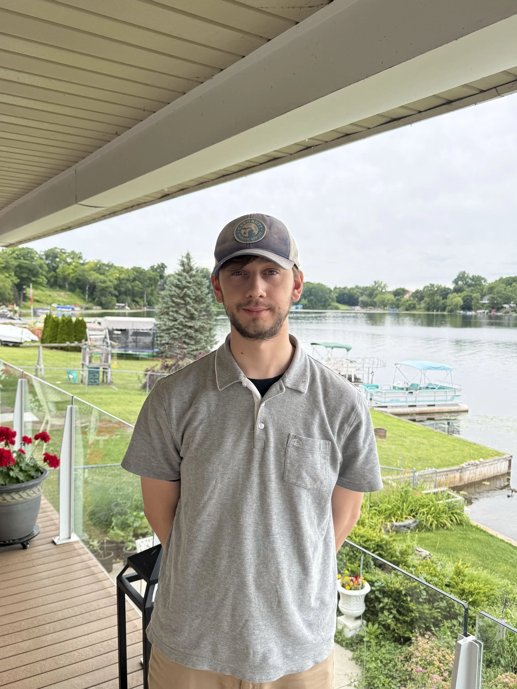 A young man with a beard and light brown hair wearing a gray cap and gray polo shirt standing on a balcony overlooking a lake with boats and green trees, under an overcast sky.