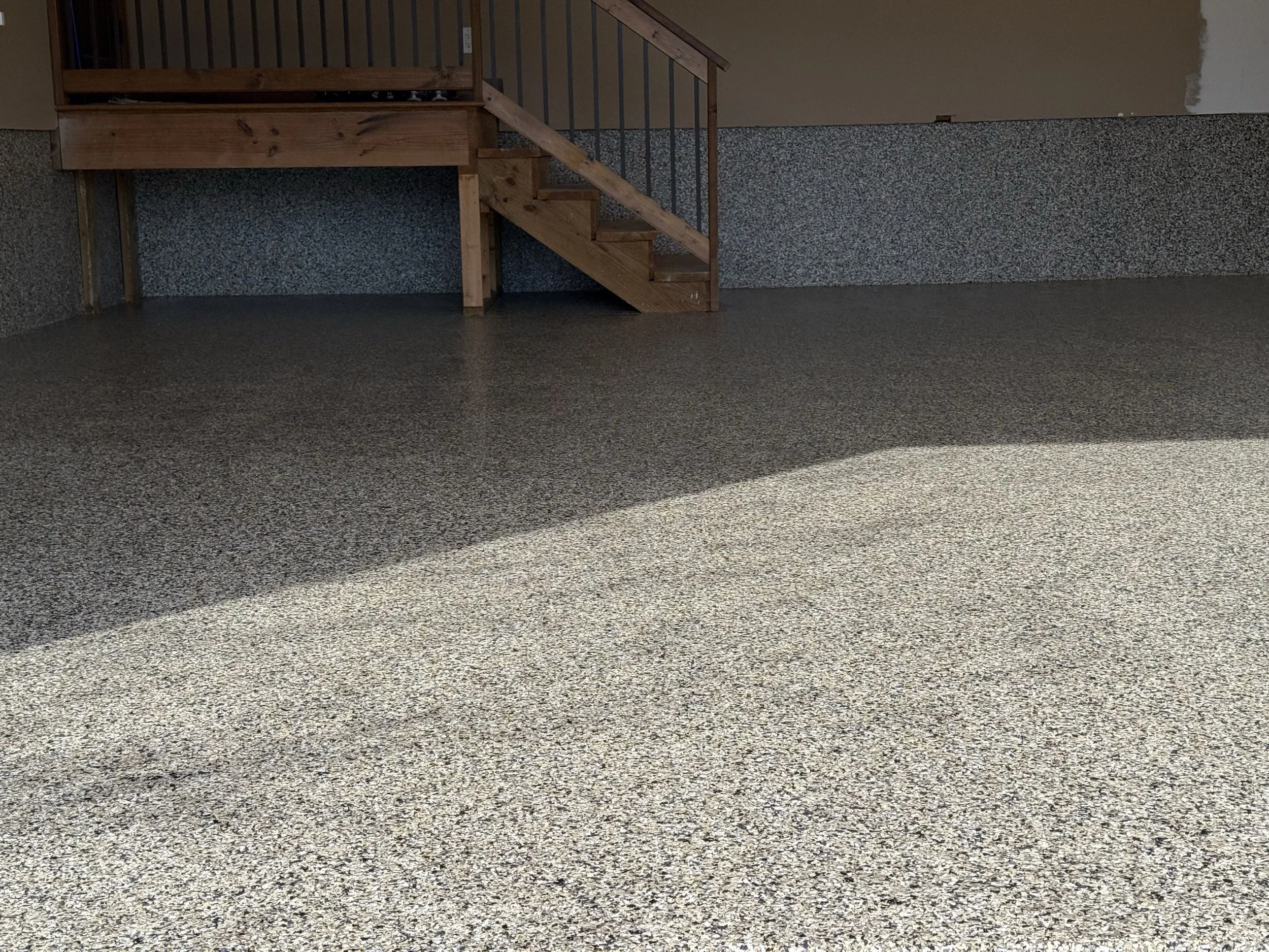Empty room with beige walls, a staircase with wooden stairs and a metal railing, and a speckled gray carpeted floor. Sunlight is streaming in, casting a shadow on the floor.