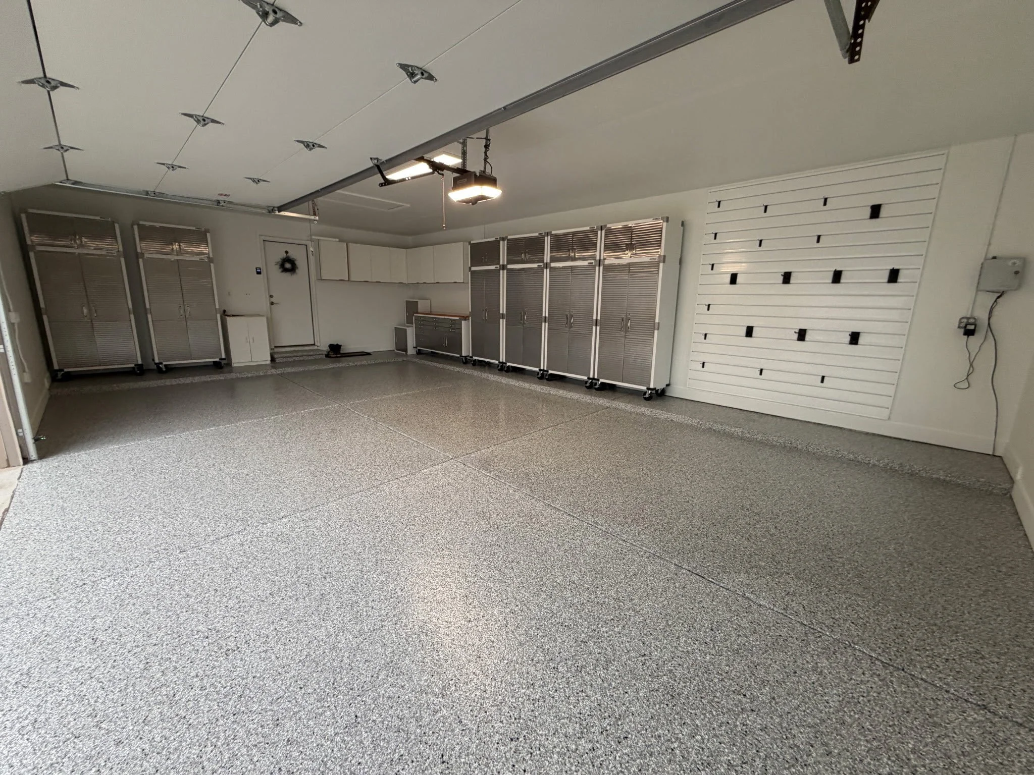 A clean garage with a textured gray floor and white walls, featuring metal storage cabinets and a decorative wreath on the door. The ceiling has a garage door opener and metal tracks.