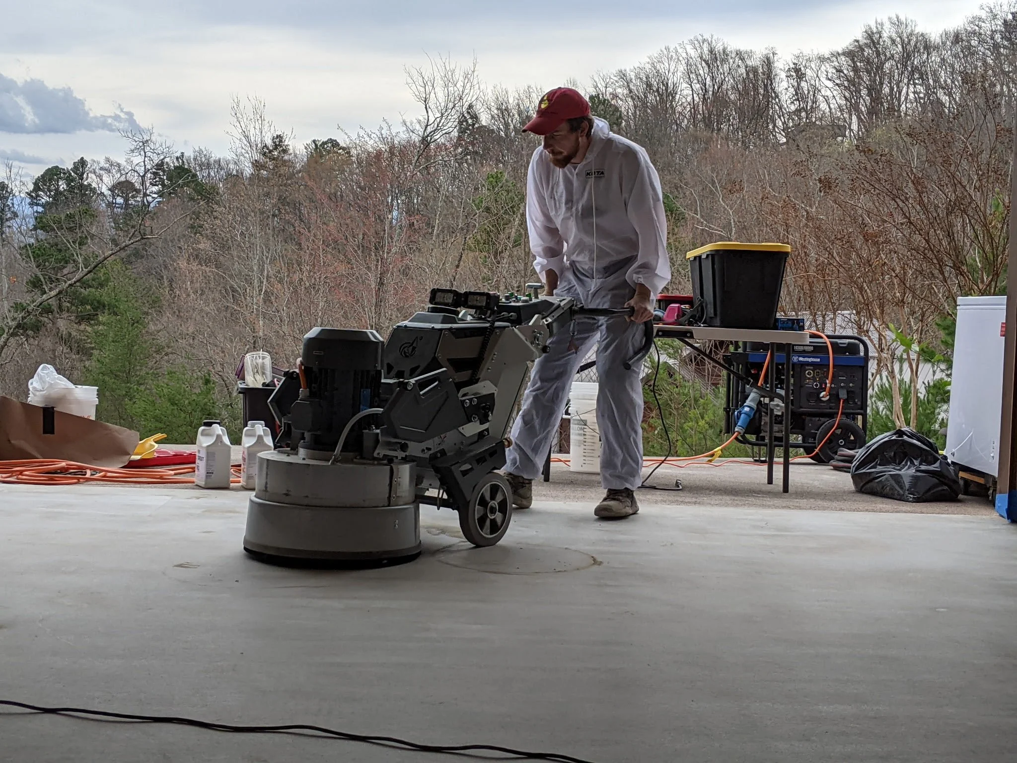 A person in white work clothes and a red cap operating a floor polishing machine outdoors, with trees in the background and tools and supplies on a table nearby.