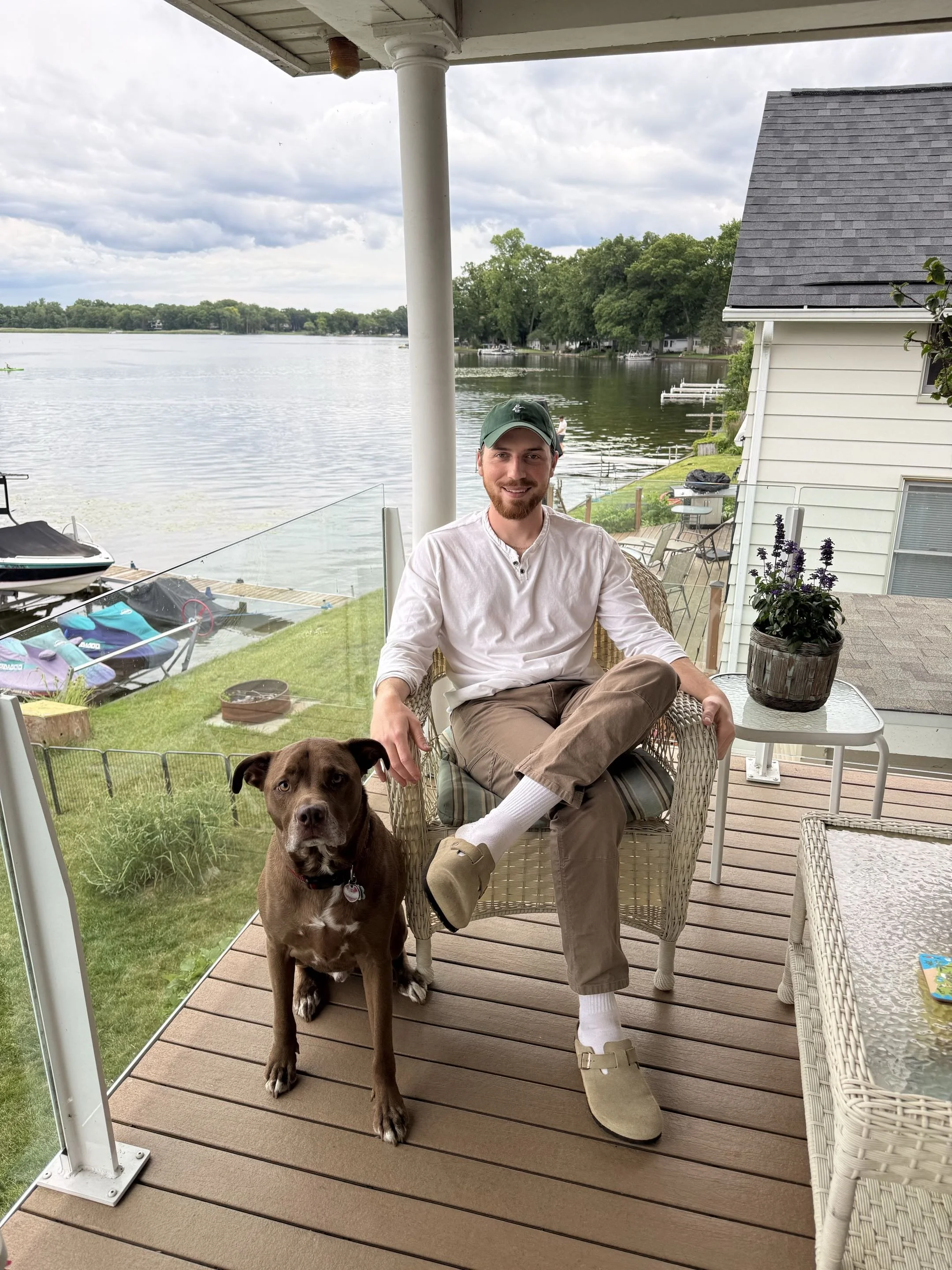 A man sitting on a chair on a porch near a lake, smiling, with a brown dog standing in front of him, and a lake view with boats and trees in the background.