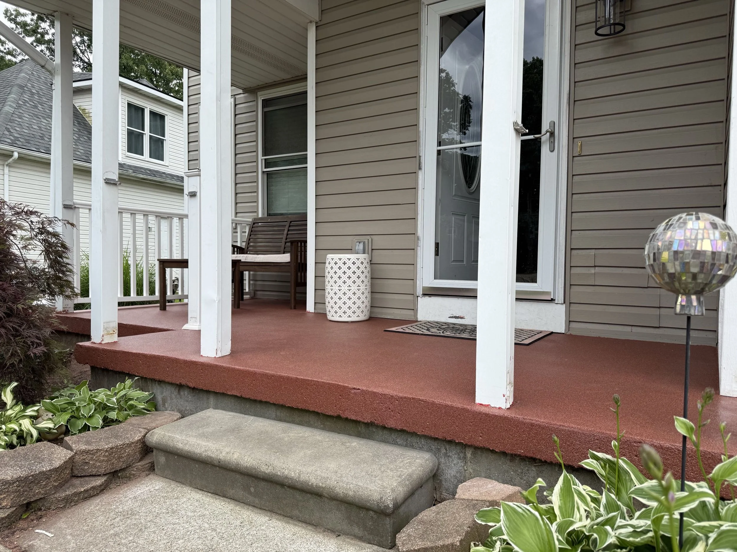 A front porch with a reddish-brown surface, white railing, a wooden bench, a decorative white lantern, a door with glass and a curtain, and a spherical garden ornament, with steps leading to a grassy area with plants.