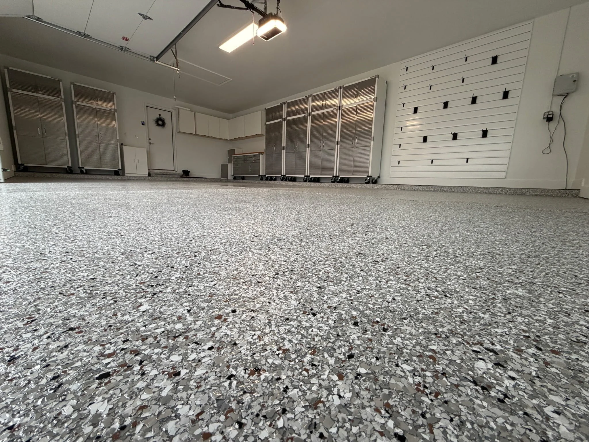 View of a garage interior with a speckled terrazzo floor, white walls, and several large silver cabinets on wheels. The ceiling has a garage door opener and a fluorescent light fixture.