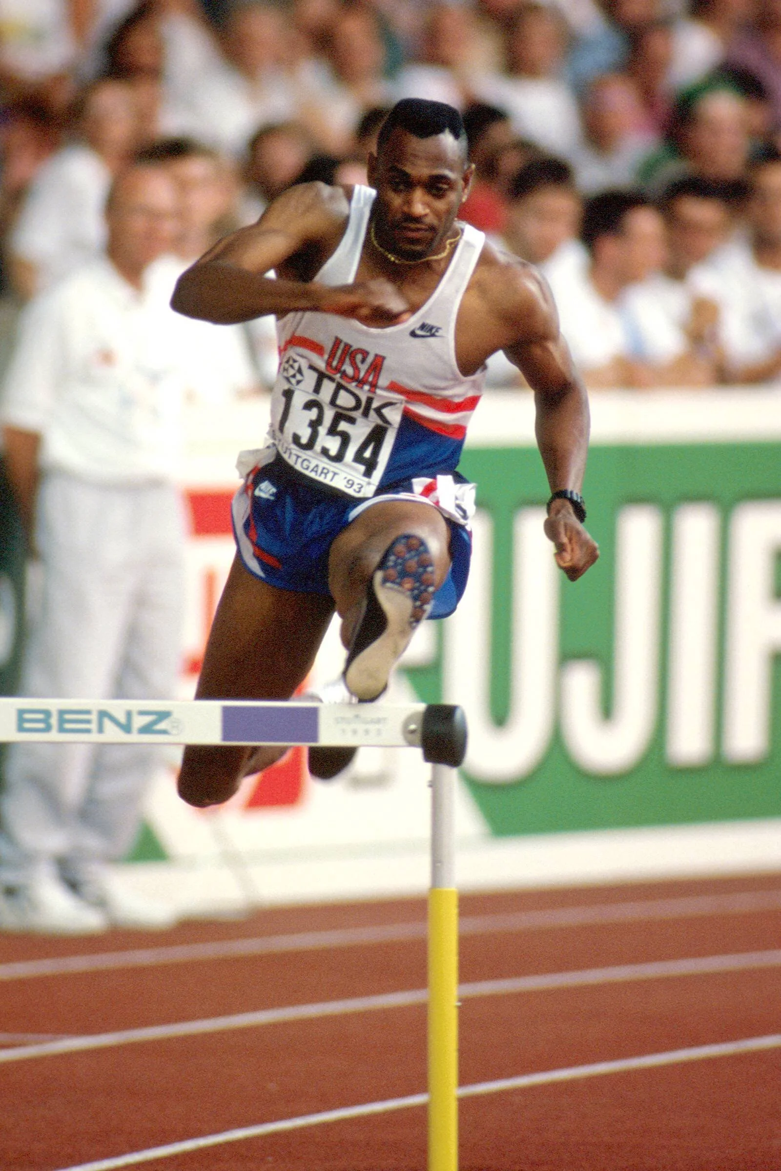 A male athlete from the USA running and jumping over a hurdle during a track and field race at an outdoor stadium. He is wearing a white and blue uniform with red accents and number 1354.